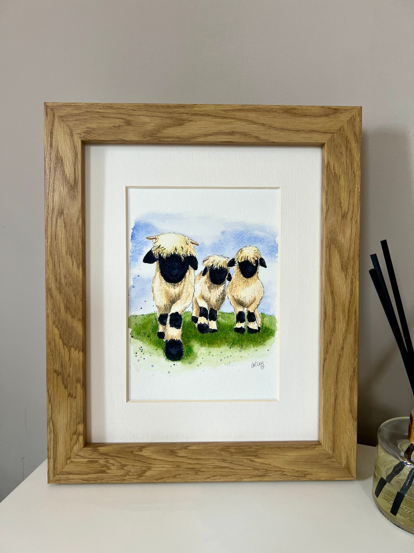A framed watercolor painting of three Valais Blacknose sheep standing in a field, with a wooden oak finish frame and an ivory-toned mount.