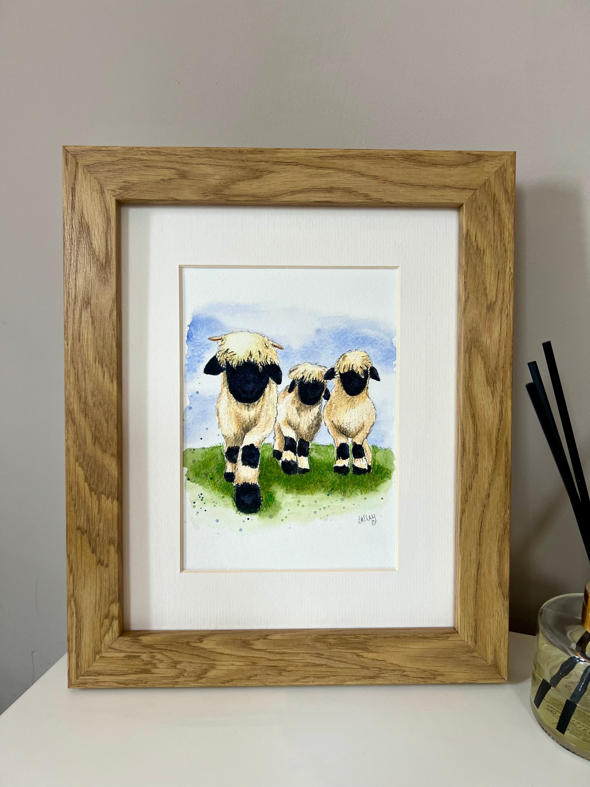 A framed watercolor painting of three Valais Blacknose sheep standing in a field, with a wooden oak finish frame and an ivory-toned mount.