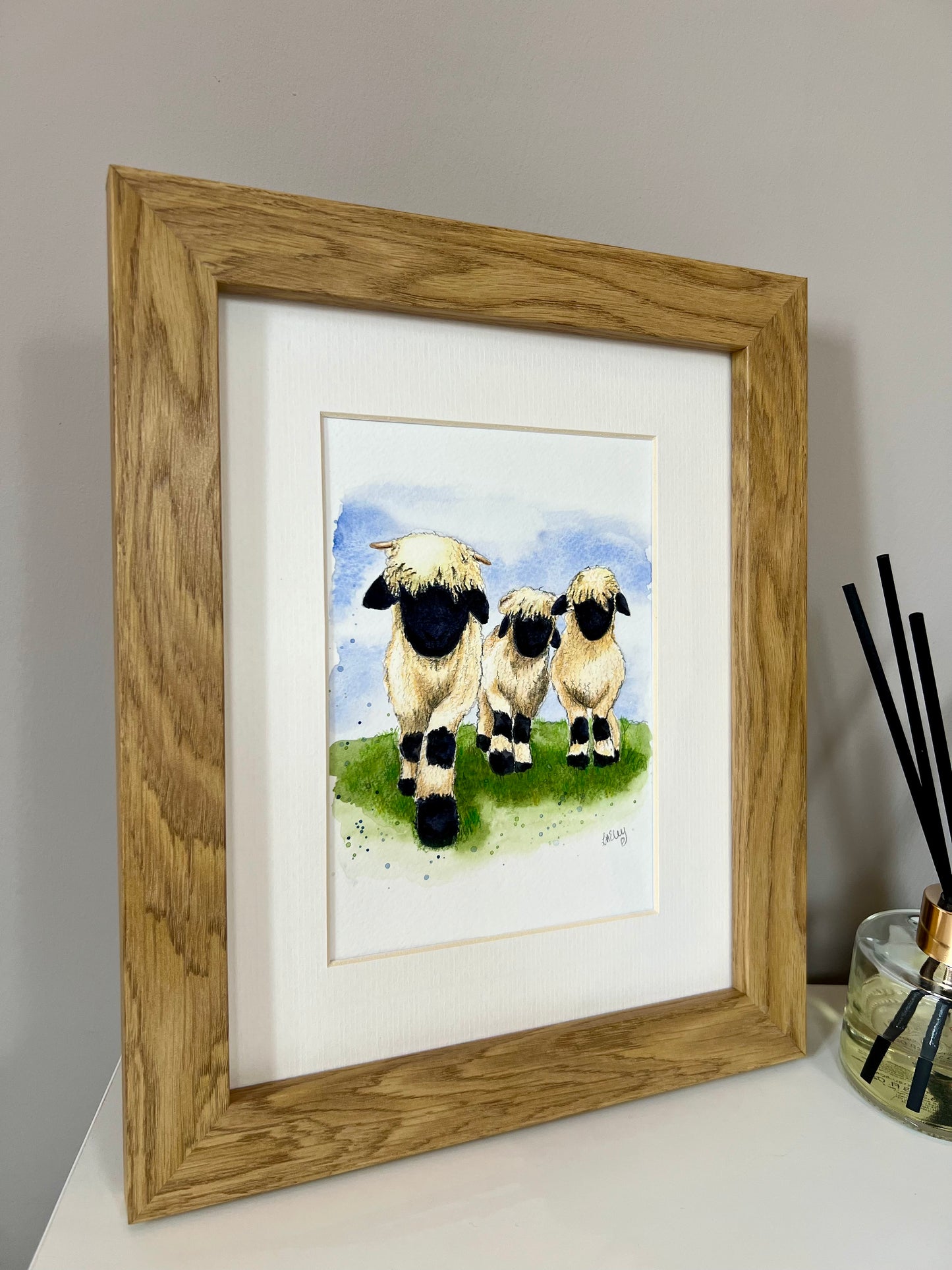 A framed watercolor painting of three Valais Blacknose sheep standing in a field, with a wooden oak finish frame and an ivory-toned mount.