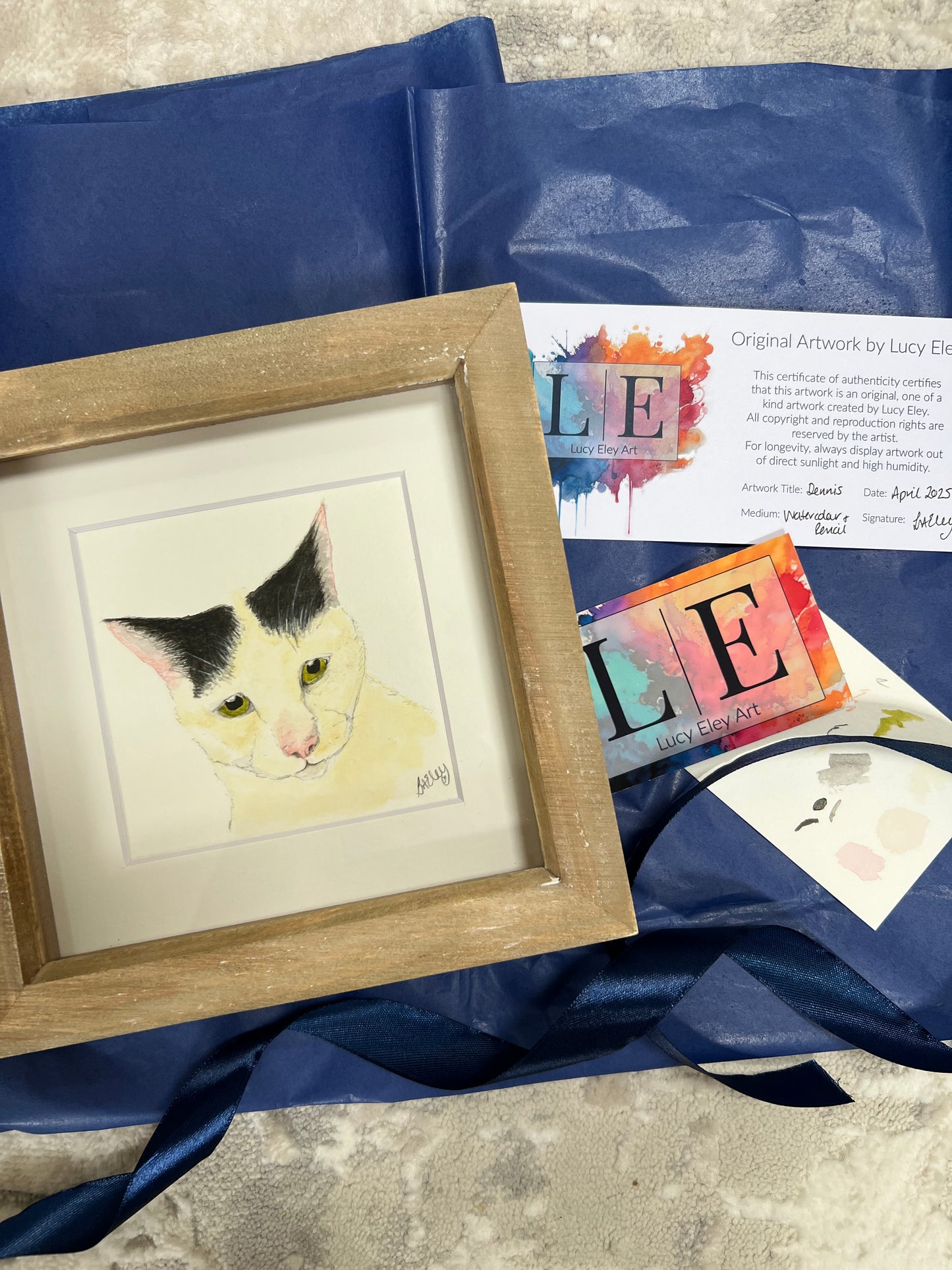 A custom watercolour pet portrait of a white cat, displayed with in a wooden frame with white mount and certificate of authenticity and wrapping paper.