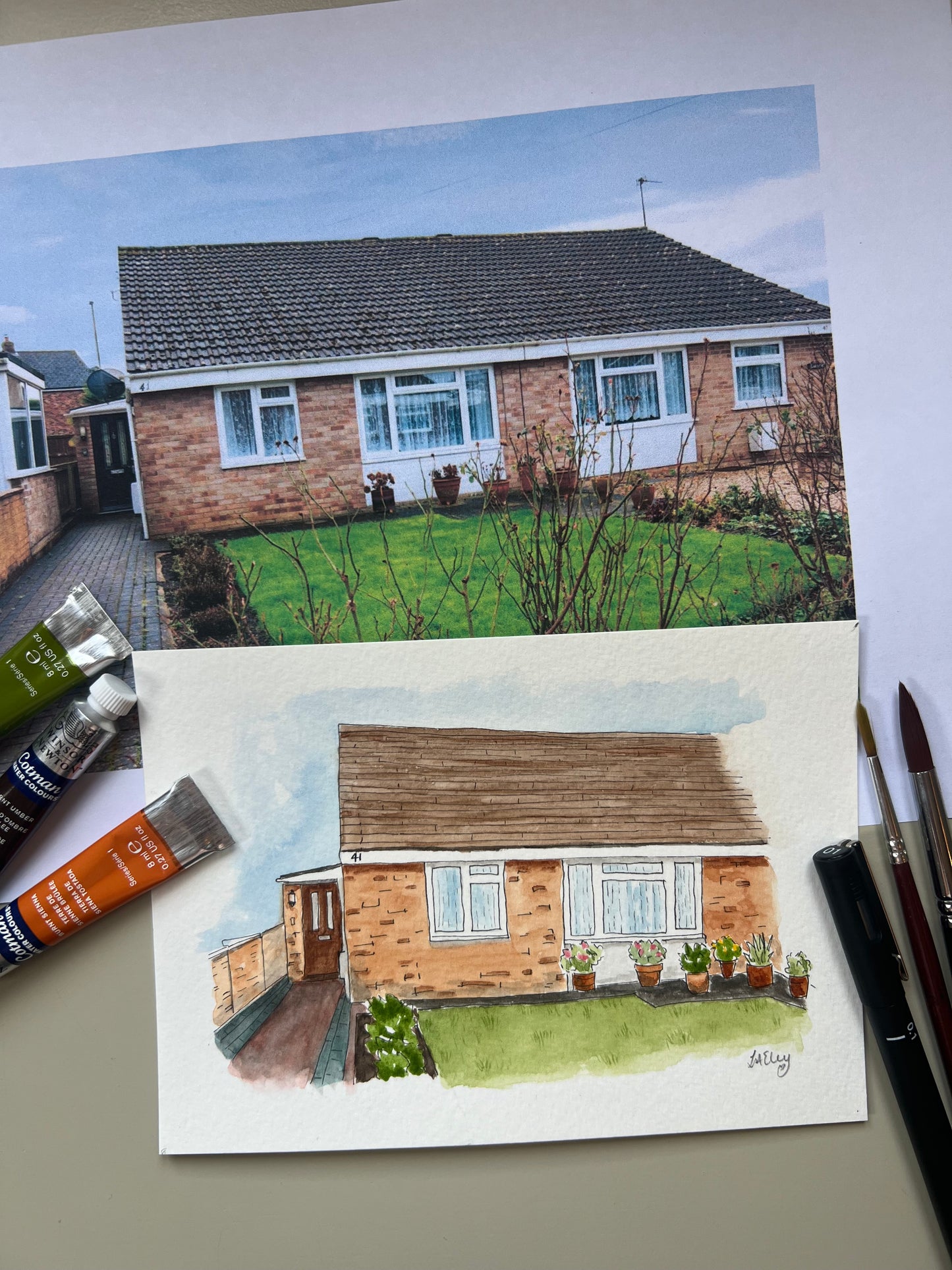 Artwork of a watercolour house portrait with paint tubes and original photo on paper in the background.