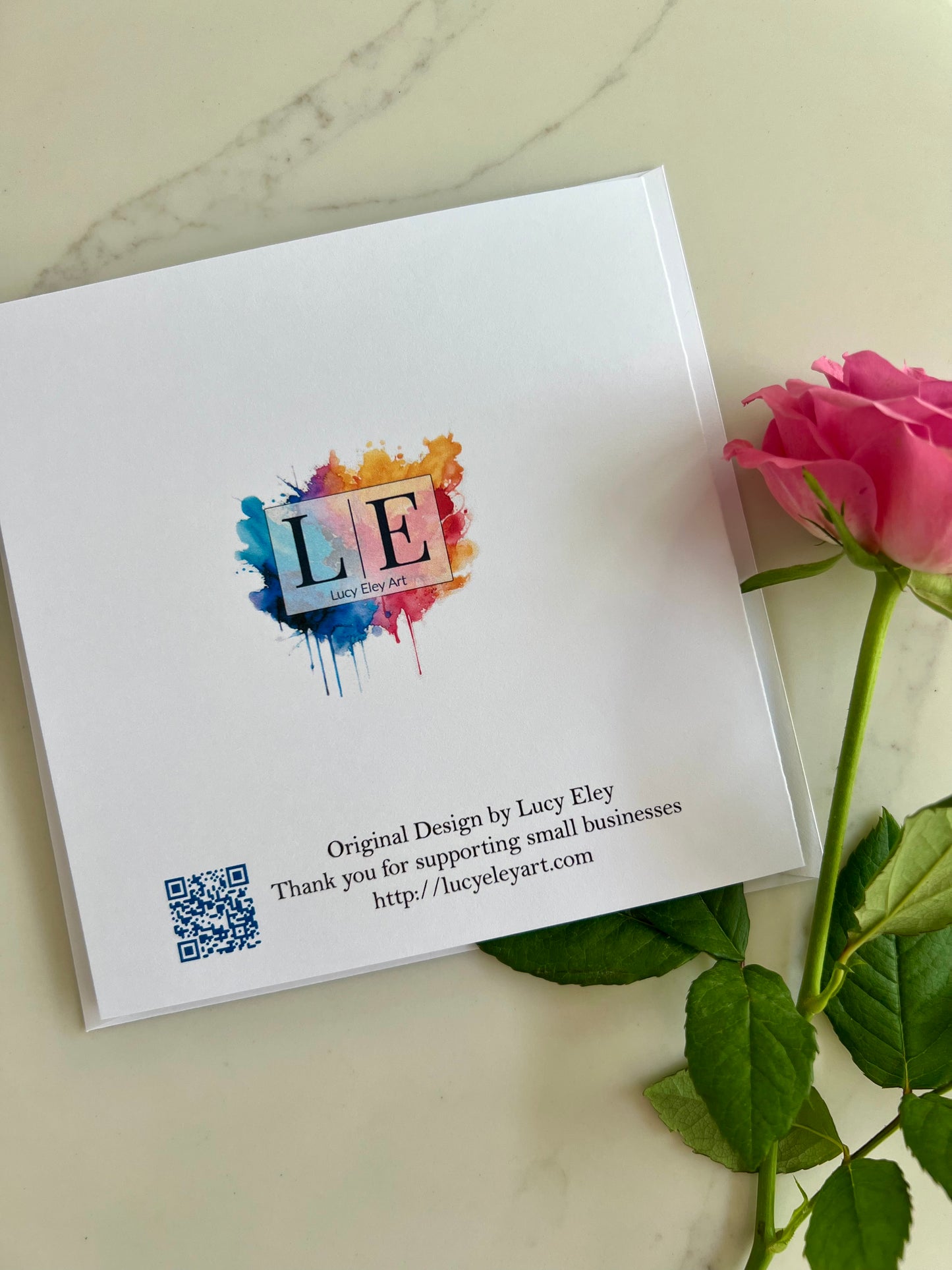 Reverse of a personalised birthday card with artist information and colorful logo and QR code on a marble surface with a pink rose.