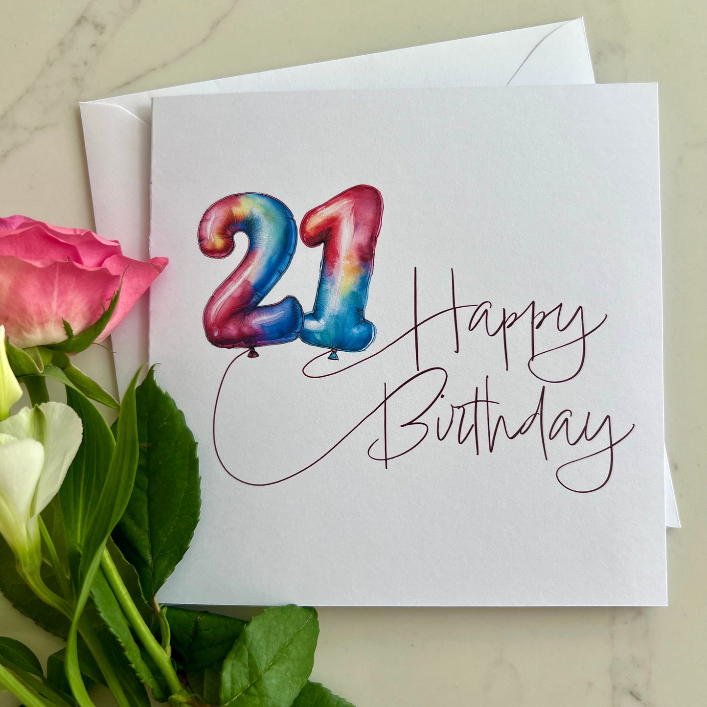 Birthday card with colorful '21' in balloons and 'Happy Birthday' text, surrounded by flowers on a marble surface.