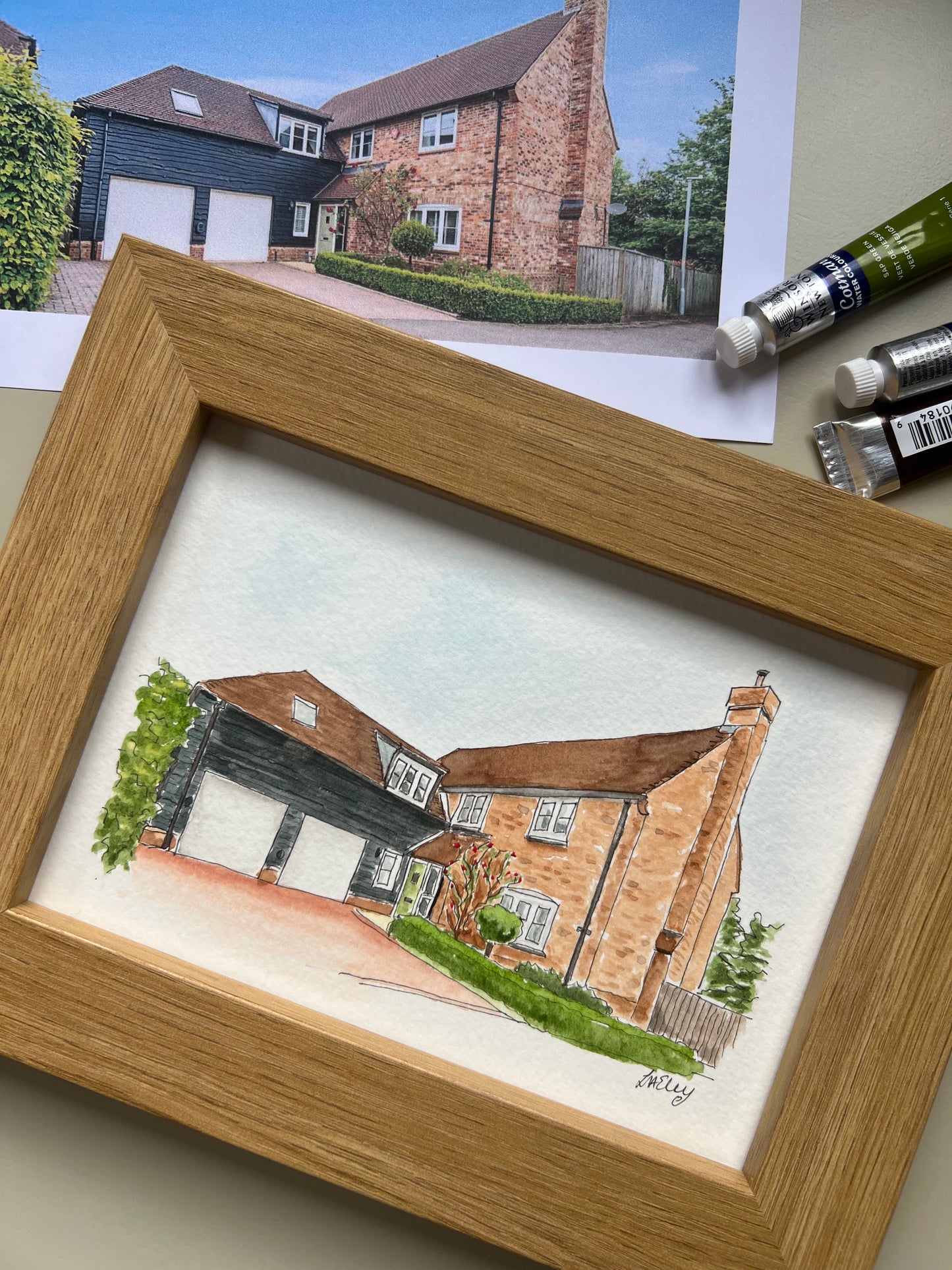 Framed drawing of a house with watercolour techniques, next to paint tubes on a light surface.