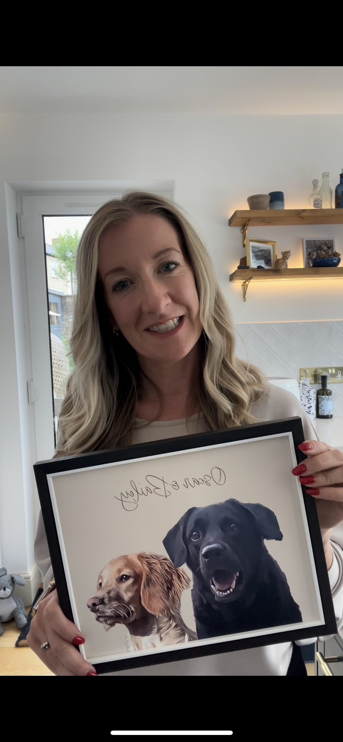 Woman artist holding a Framed portrait artwork of two dogs, one brown spaniel and one black retriever in a black frame in a kitchen setting.