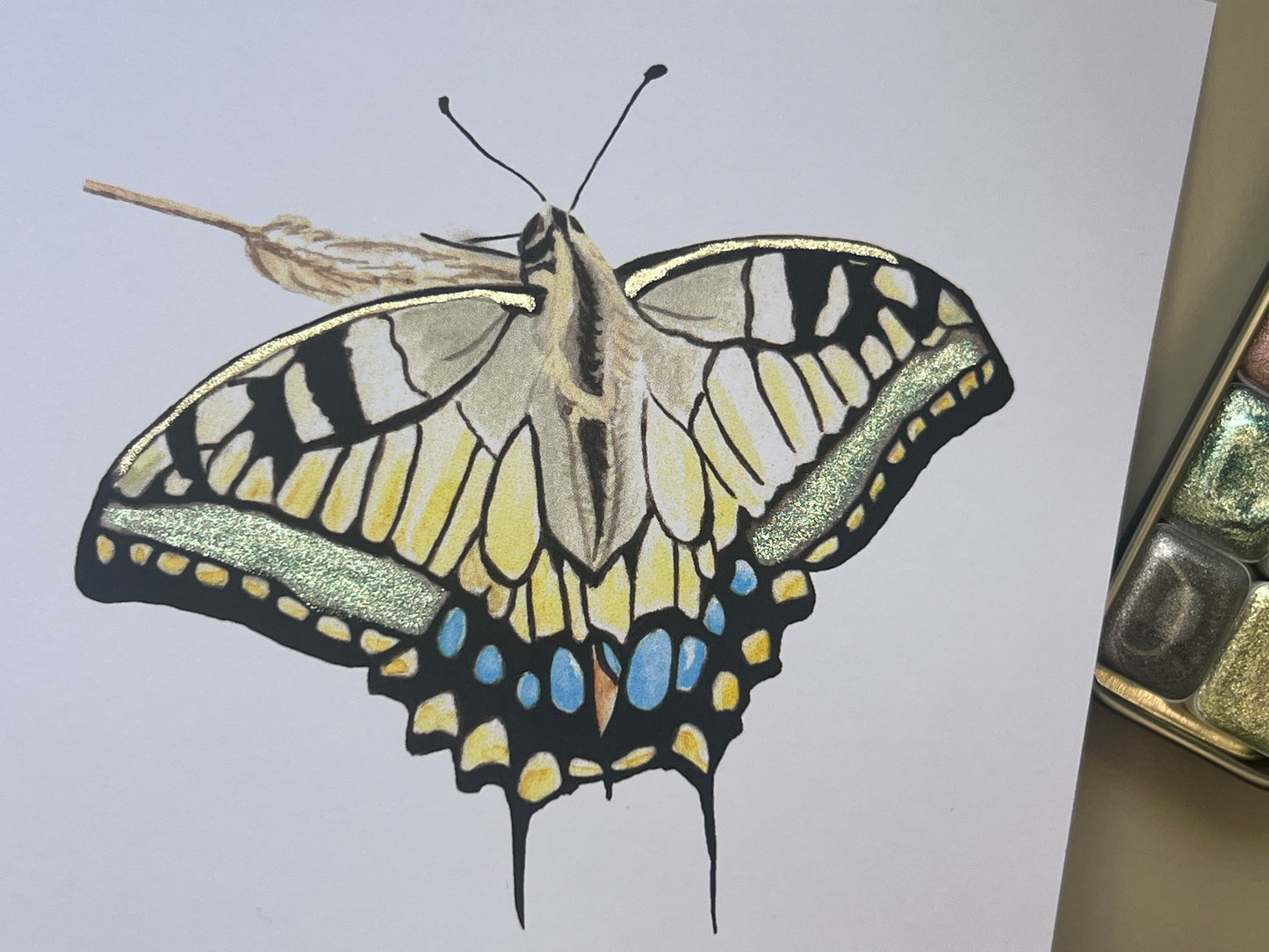 Swallowtail Butterfly illustration with green and gold glittery metallic finish on a white card in painting studio.