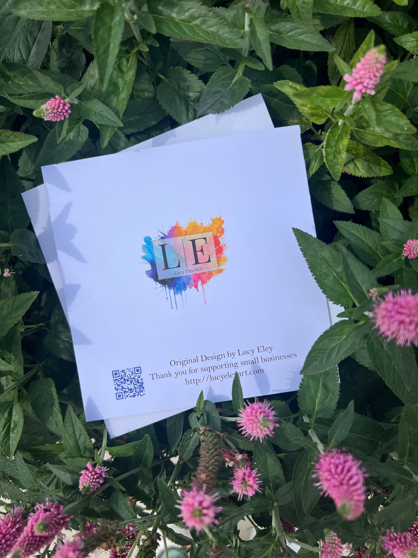 Reverse of handmade card with colorful logo and text on a bed of green leaves and pink flowers