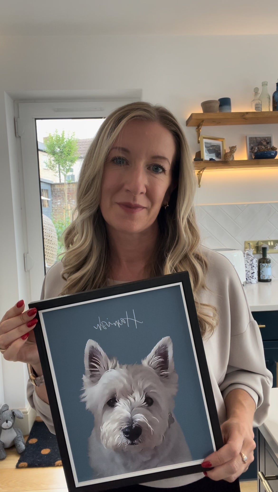 Woman artist holding a framed custom portrait of a white westie dog with a blue background in a kitchen.