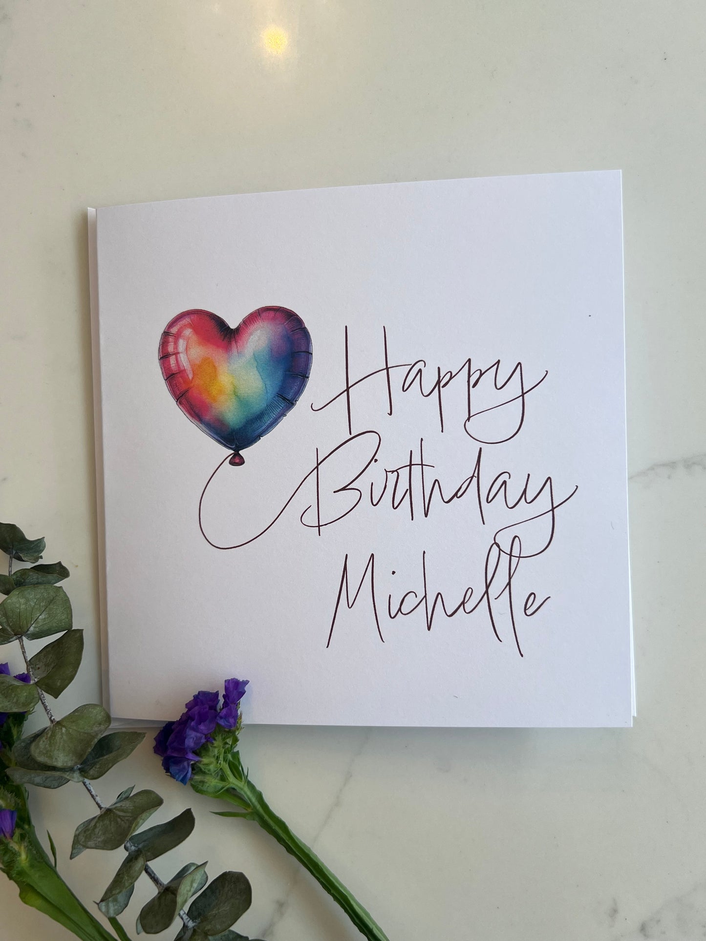 Birthday card with a heart-shaped balloon and personalized name on a marble surface with flowers.