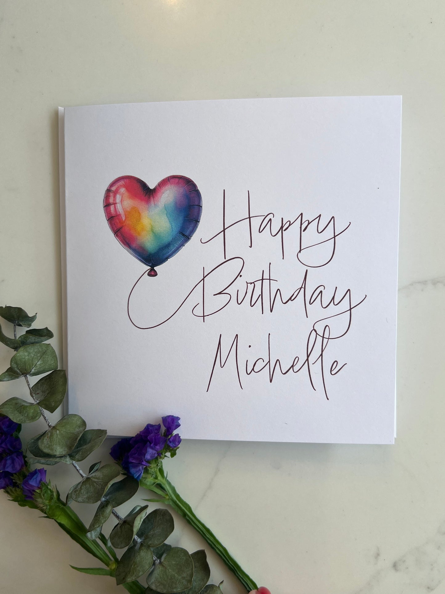 Birthday card with a heart-shaped balloon and personalized name on a marble surface with flowers.