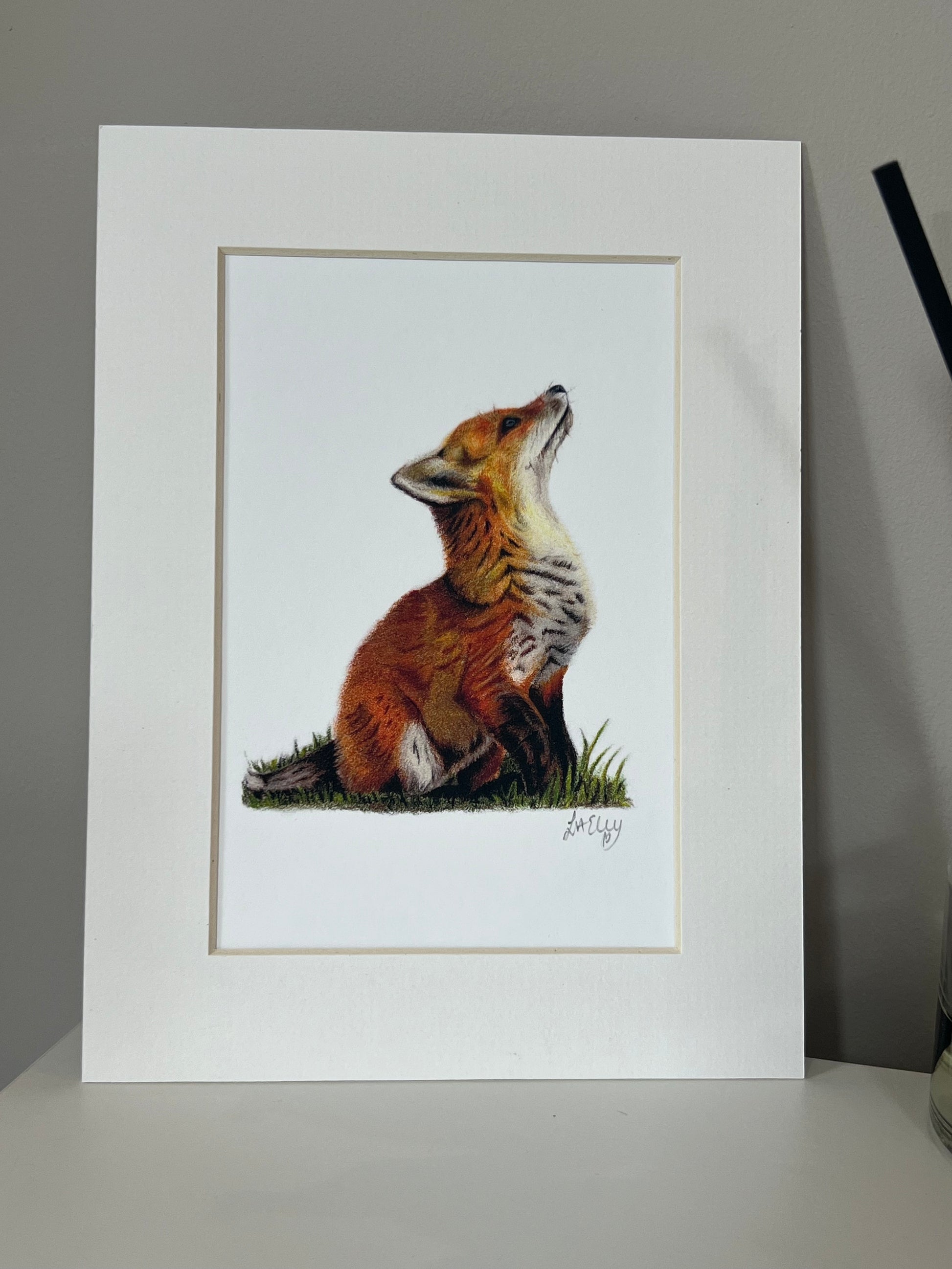 Artwork of a fox cub in vibrant oranges and yellows on a white background in a white mount.