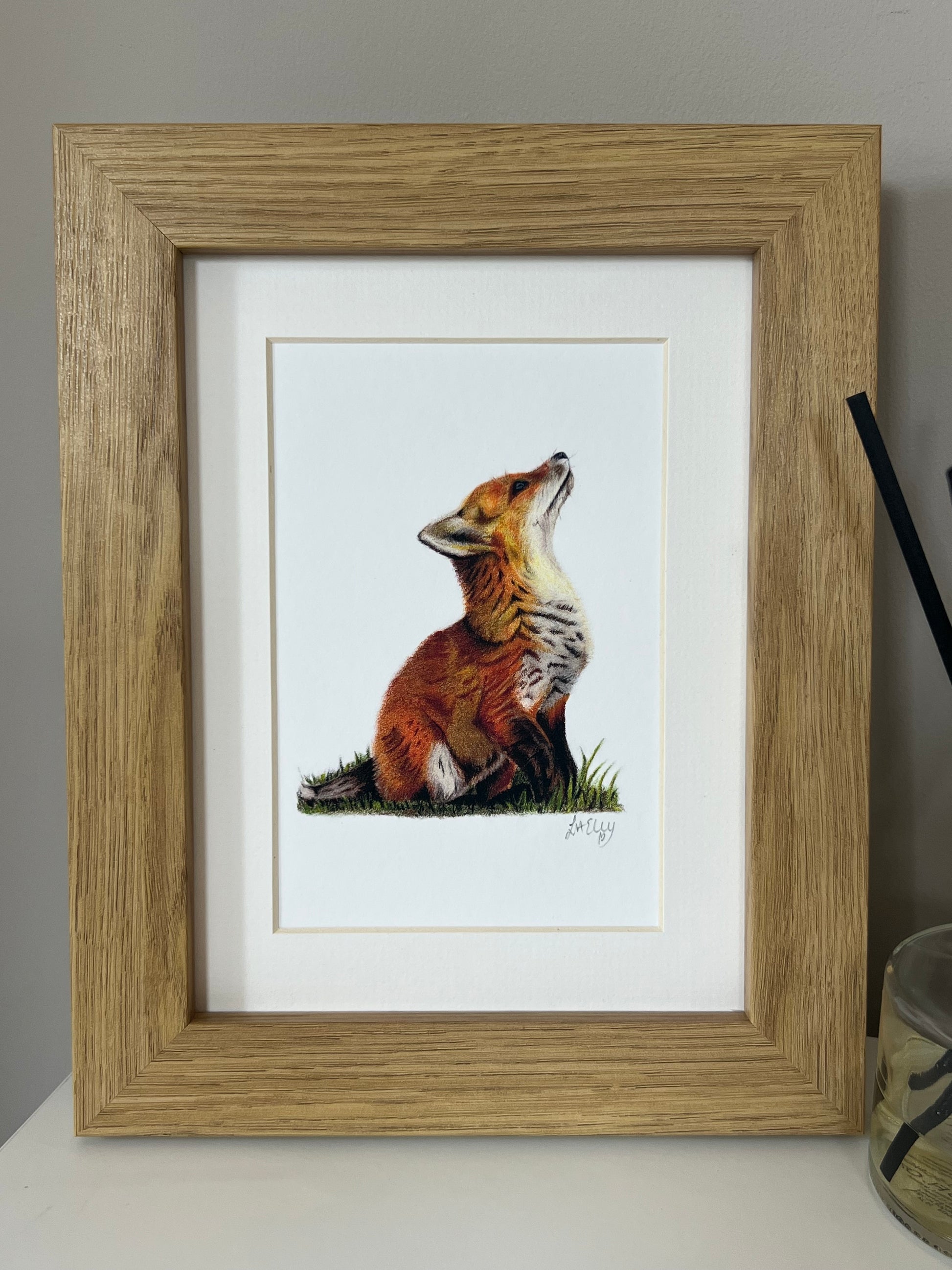 Framed artwork of a fox cub in vibrant oranges and yellows on a white background in a wooden oak style frame.