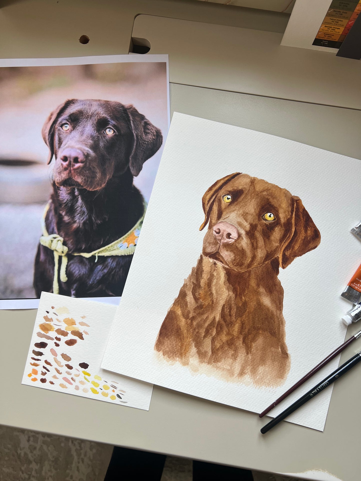 A custom watercolour portrait of a chocolate brown labrador dog, with paint and pencil supplies.