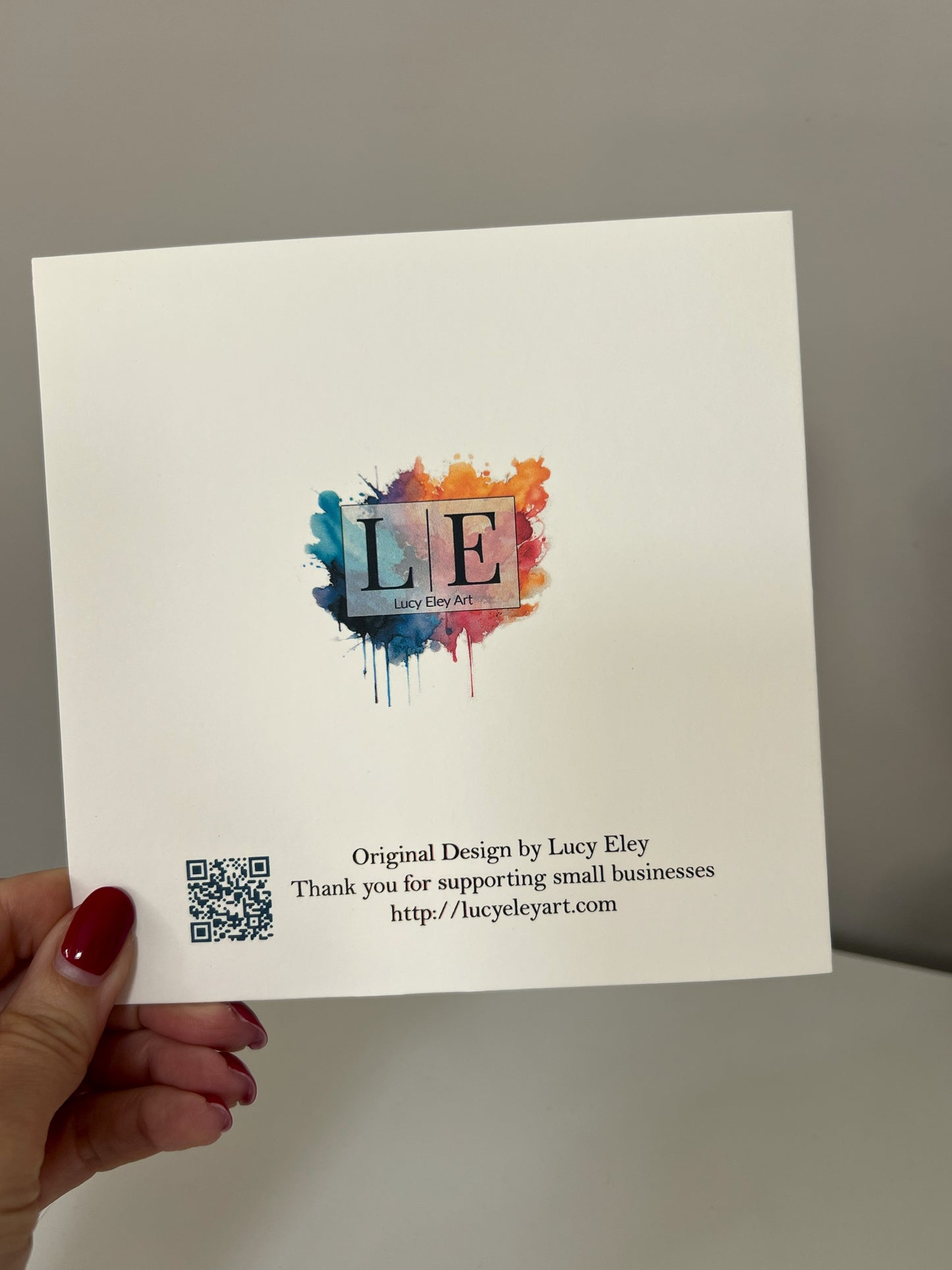 A hand with red painted nails holds a white square card featuring a colorful paint-splatter logo with the letters LE and text reading 'Original Design by Lucy Eley' plus a QR code, against a plain gray background.