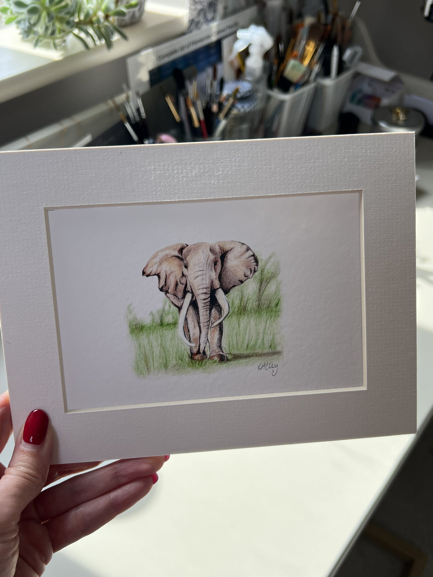 A hand with red nail polish holds a matted art print featuring a pencil illustration of an elephant standing in grass. The print has a wide white border and is photographed in a real indoor setting with a desk and art supplies blurred in the background.