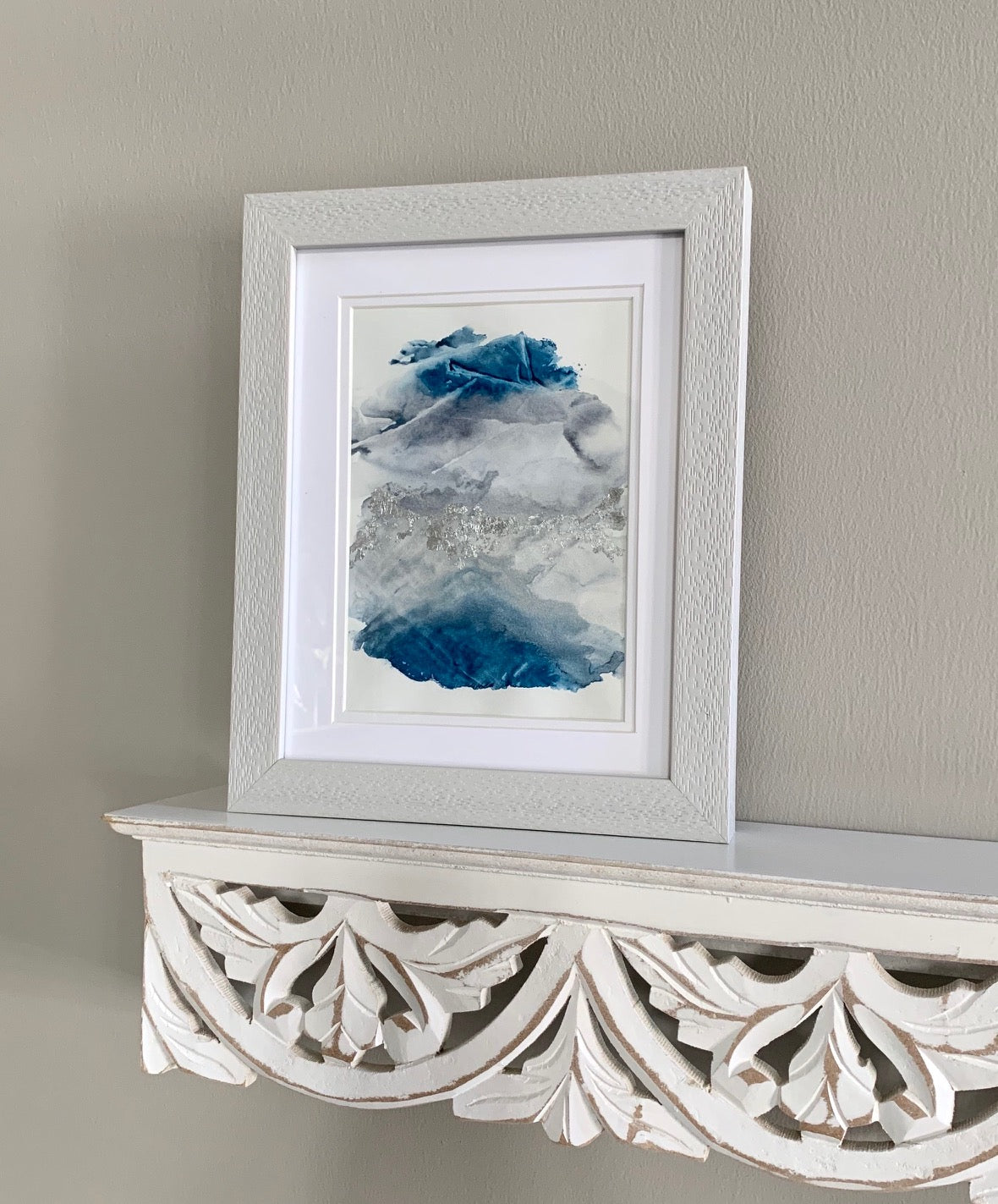 A framed abstract watercolor painting with blue and grey tones, displayed on a white ornate shelf.