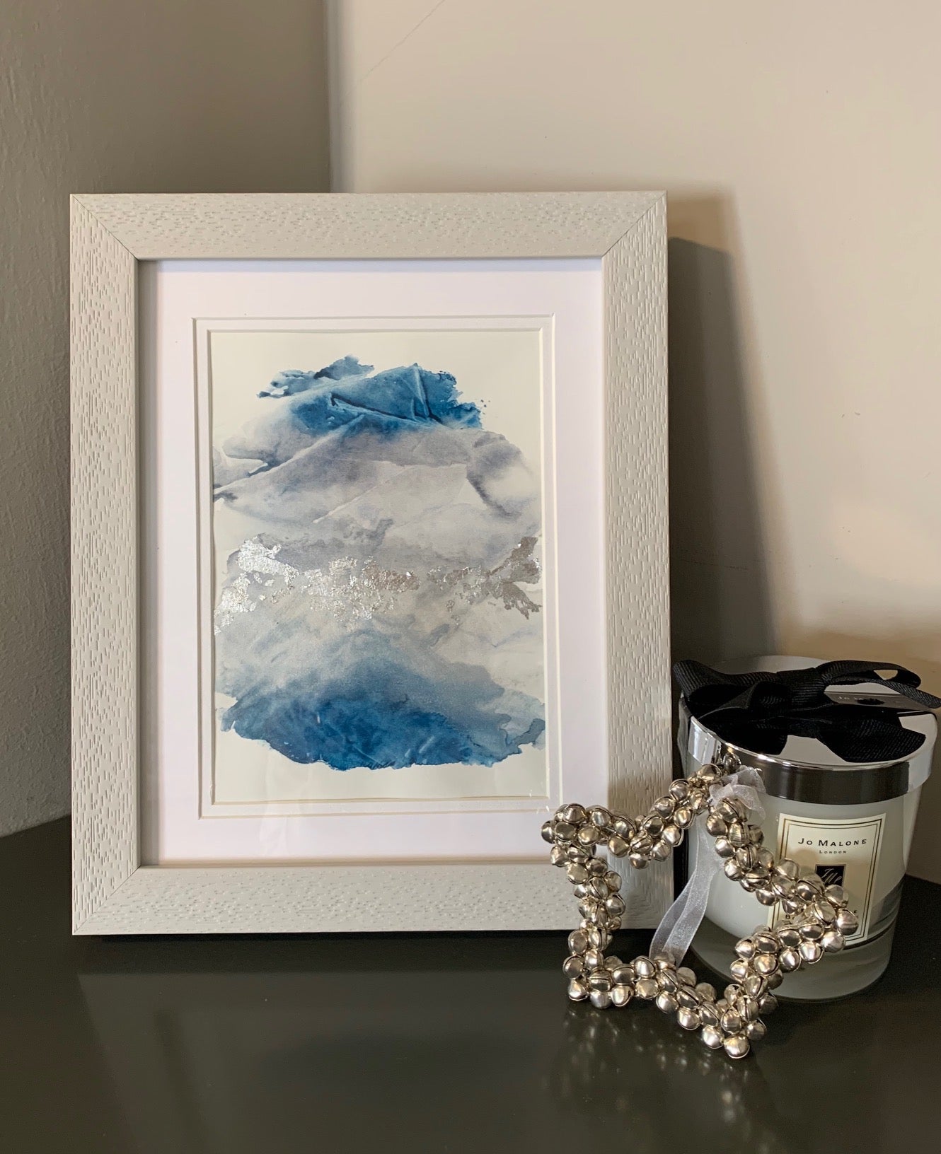 A framed abstract watercolor painting with blue and grey tones, displayed on a grey shelf.