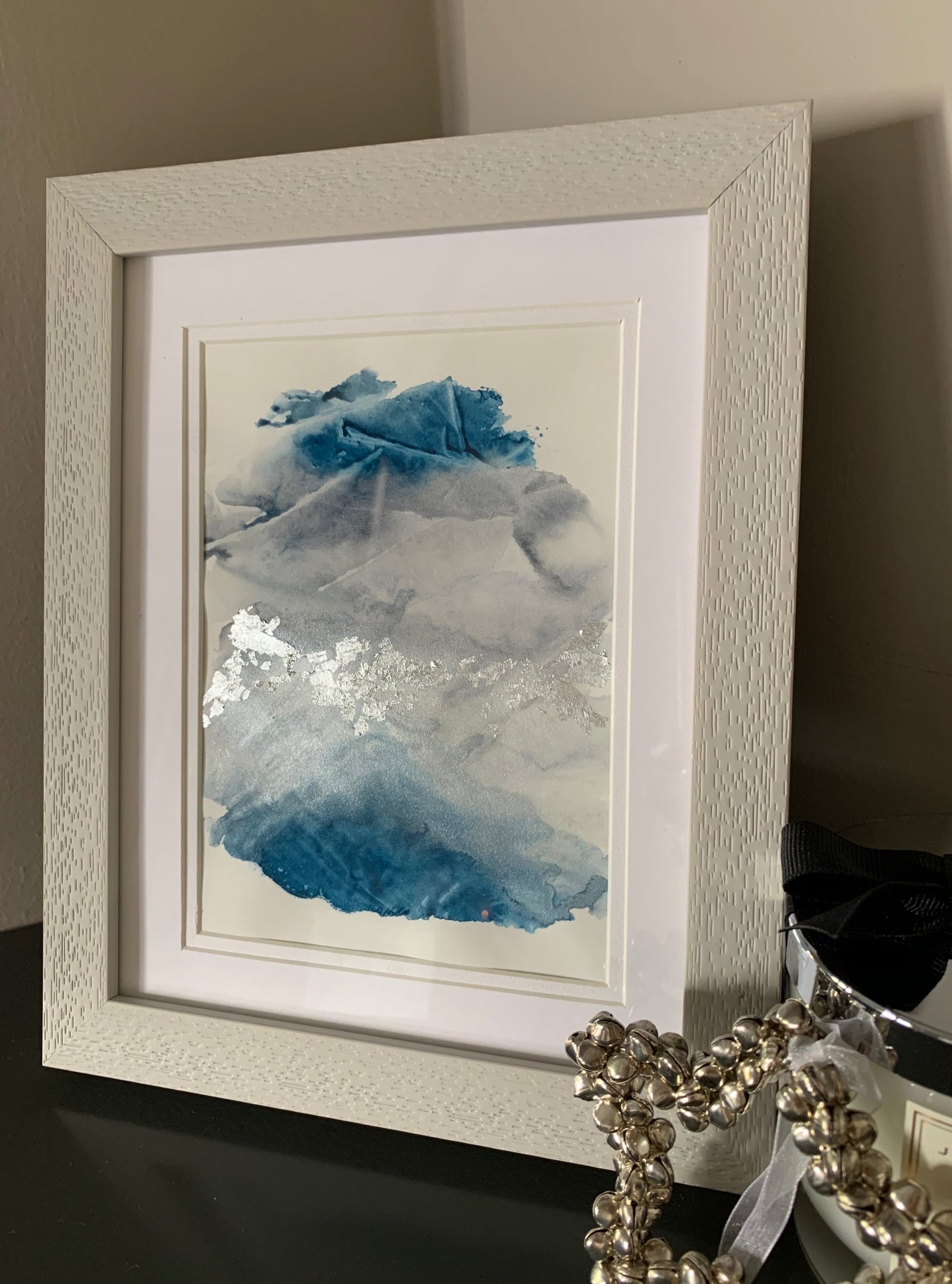 Close up of a framed abstract watercolor painting with blue and grey tones, displayed on a grey shelf.