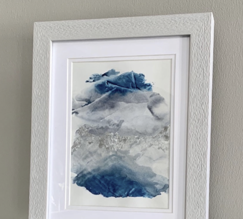 Close up of a framed abstract watercolor painting with blue and grey tones, displayed on a grey shelf.