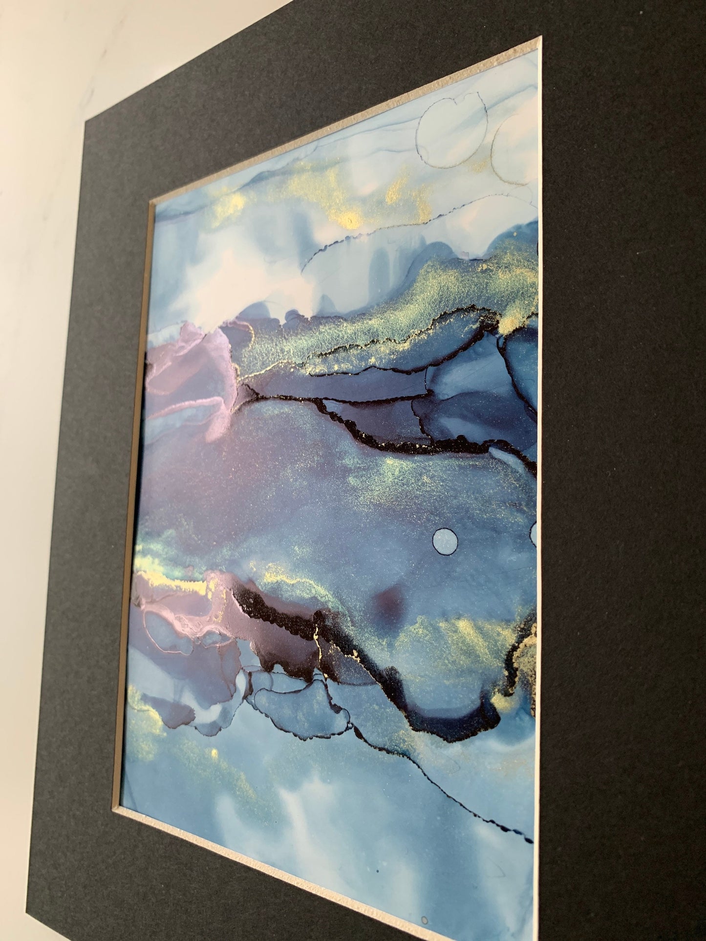 An A5 size contemporary ink painting featuring blue and metallic colours with gold shimmers, presented in a black mount.