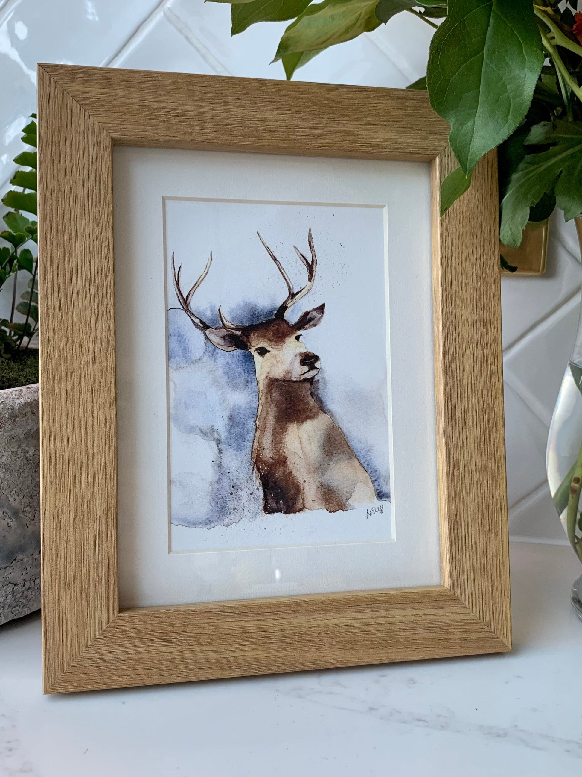 Watercolour stag in brown, grey and blue tones in a white mount and wooden frame.