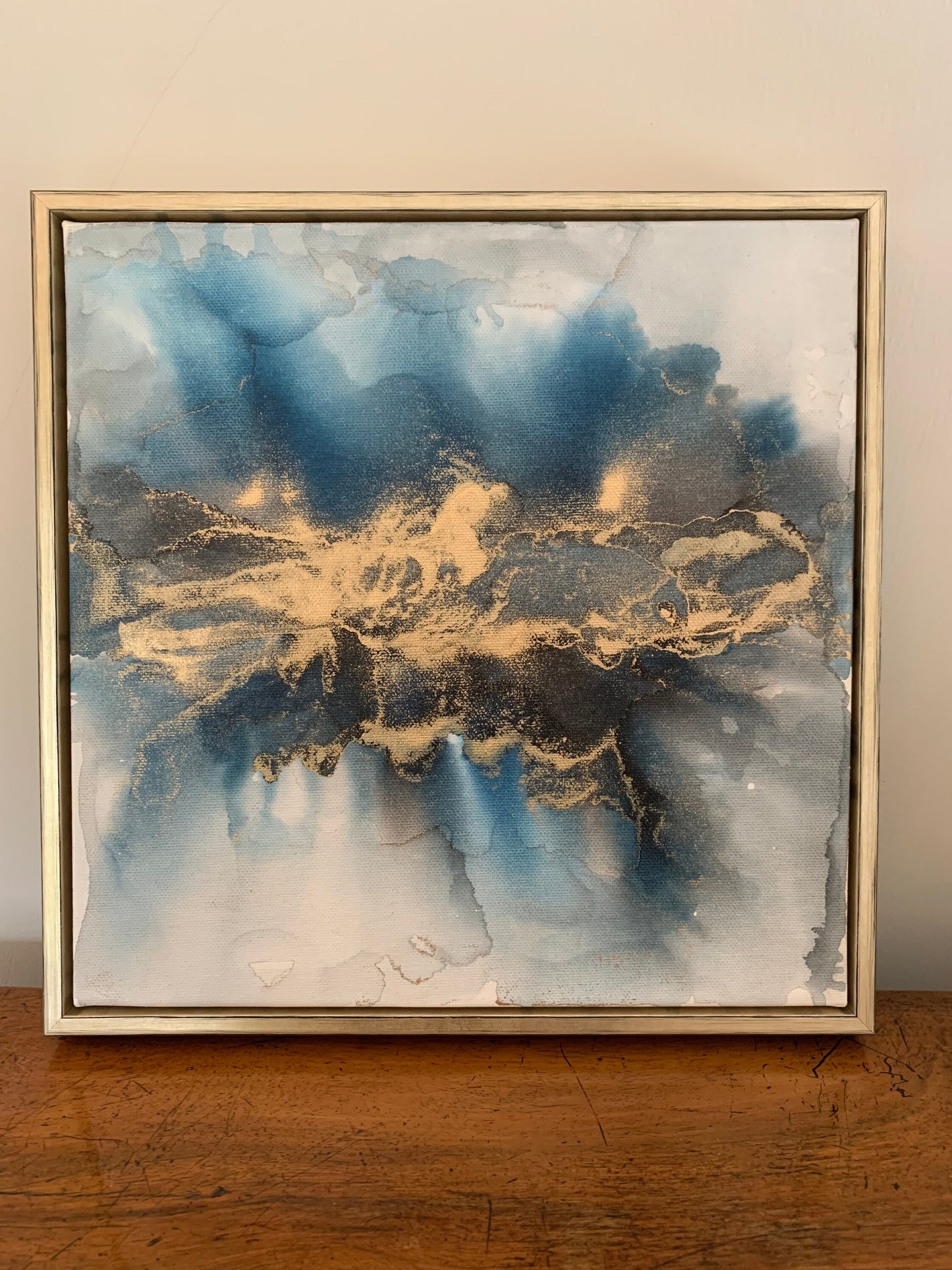 A one-off statement canvas painting featuring an abstract design in alcohol inks with gold and blue colours, with a metallic finish in a gold box frame.