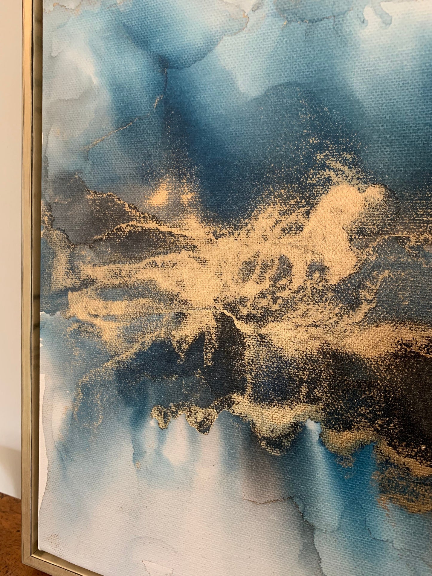 Close up of a one-off statement canvas painting featuring an abstract design in alcohol inks with gold and blue colours, with a metallic finish in a gold box frame.