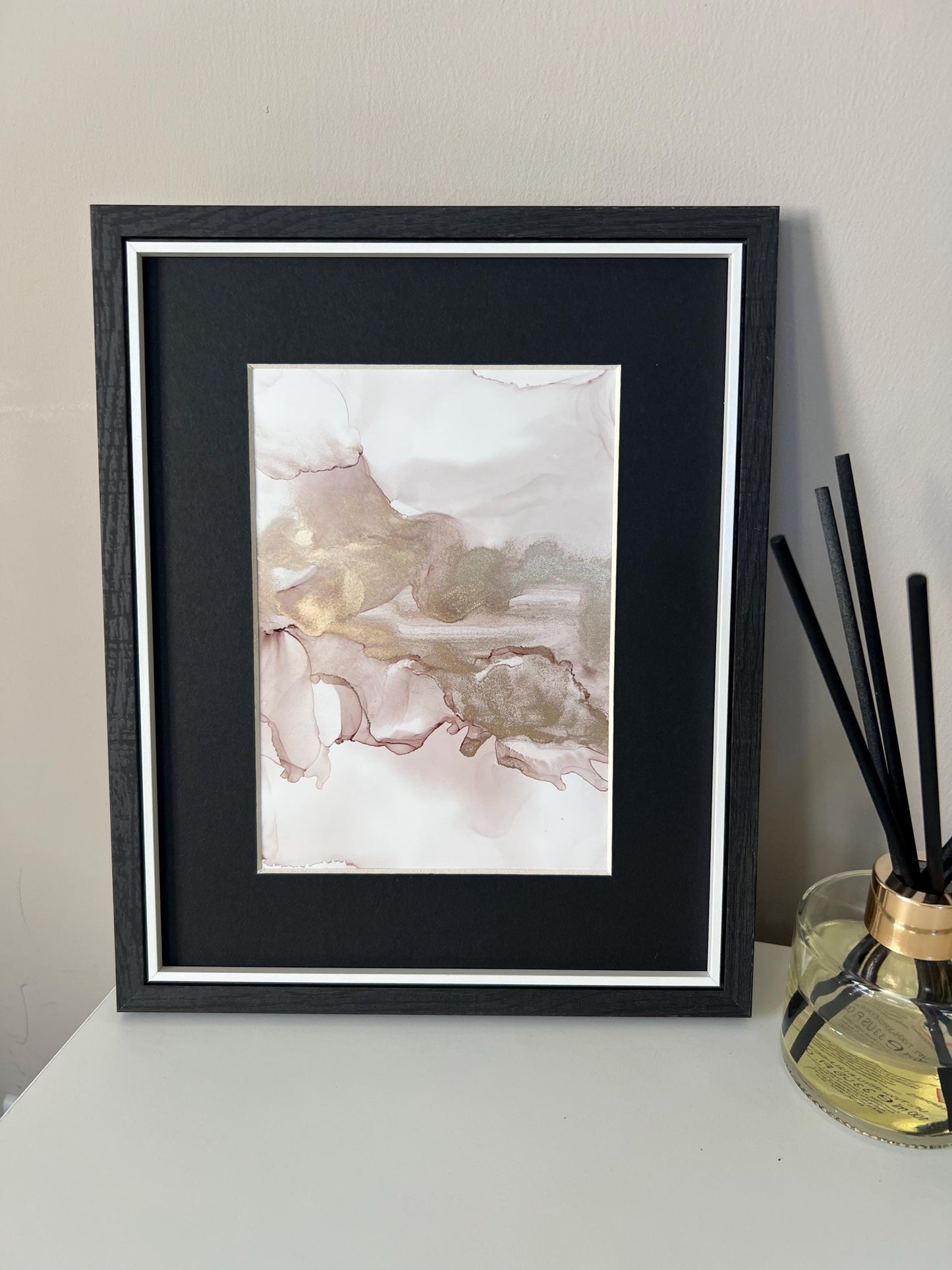 An A5 size contemporary ink painting with pink and gold metallic colors, presented in a black frame, placed on a white surface next to decorative items.