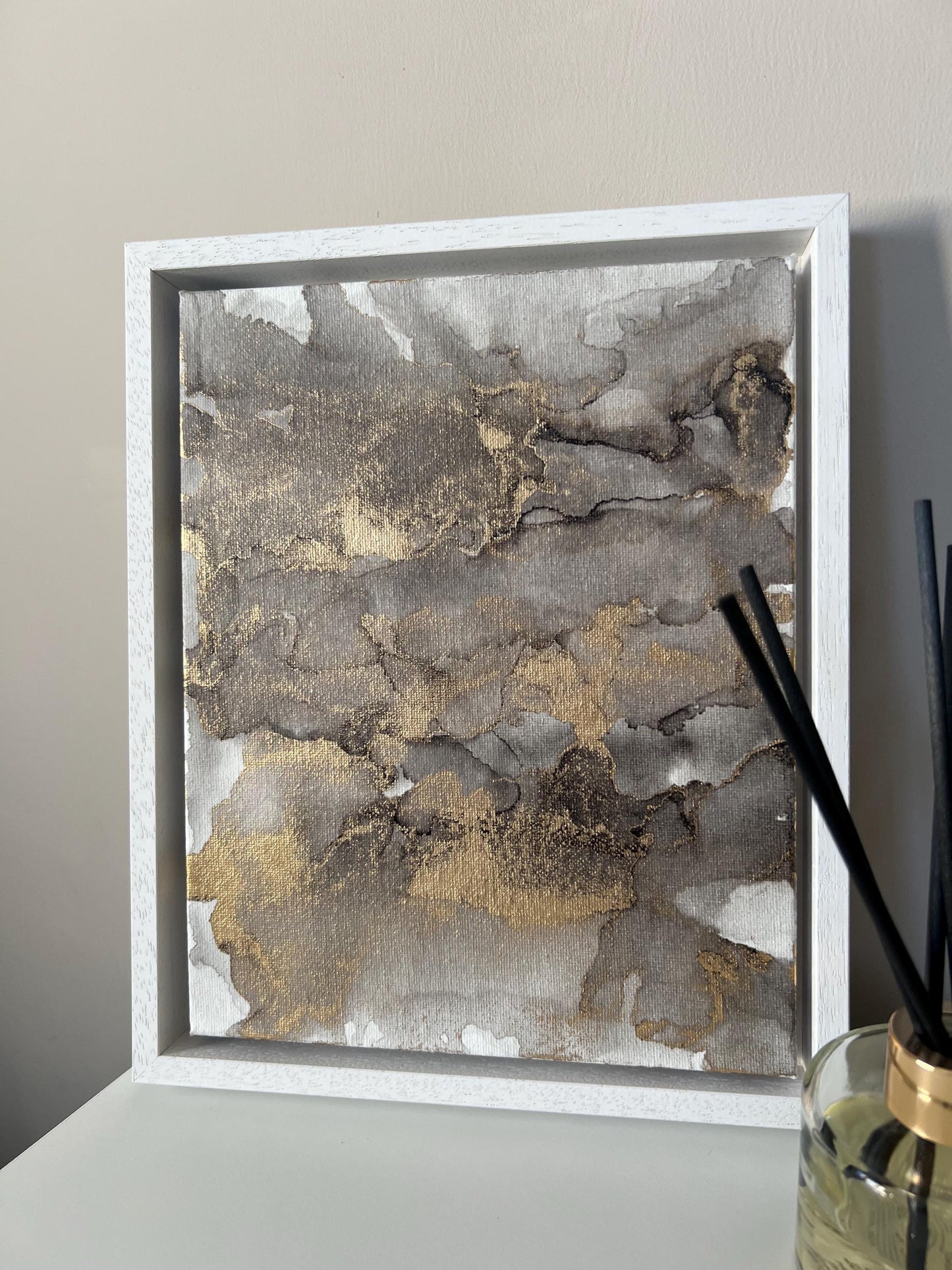 An abstract grey black and gold alcohol ink painting on canvas with a white frame, displayed against a grey background.