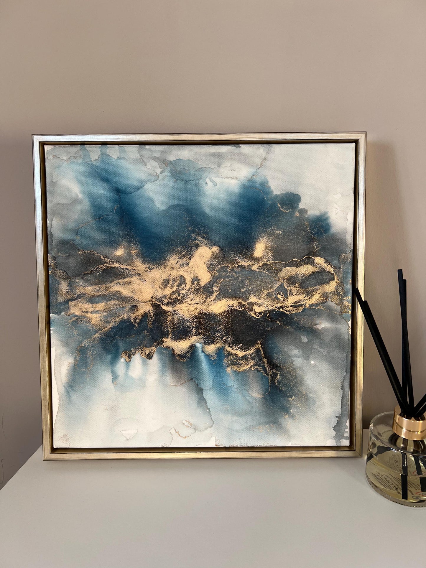 A one-off statement canvas painting featuring an abstract design in alcohol inks with gold and blue colours, with a metallic finish and in a gold finish box frame.