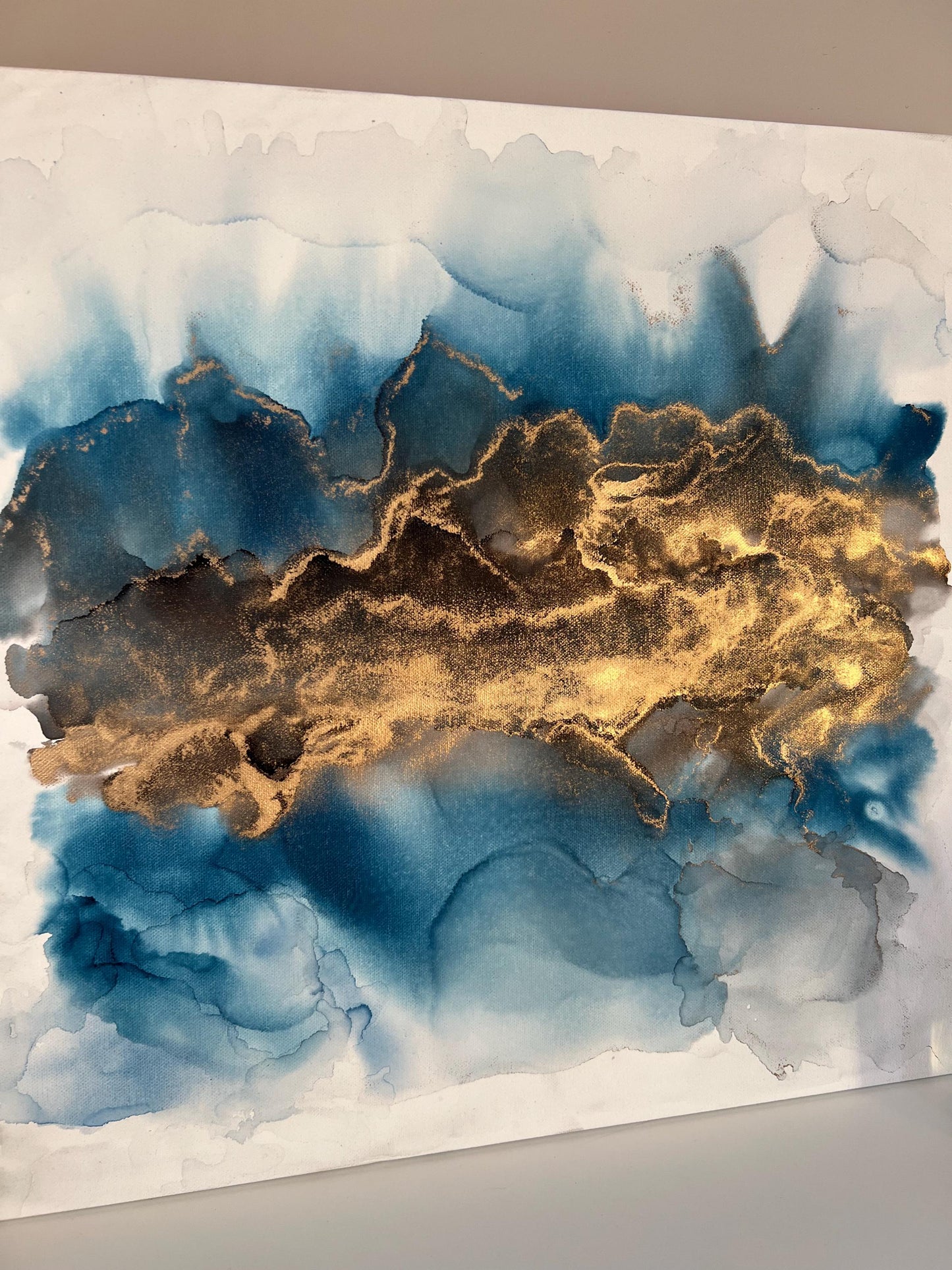 A one-off statement canvas painting featuring an abstract fluid motion design in alcohol inks with gold and blue colours, with a metallic finish.