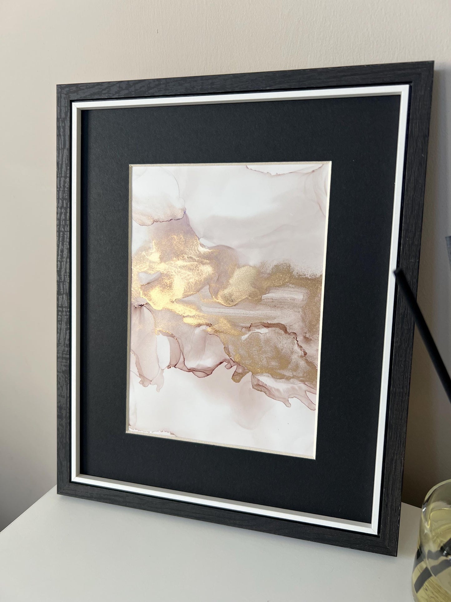 An A5 size contemporary ink painting with pink and gold metallic colors, presented in a black frame, placed on a white surface next to decorative items.