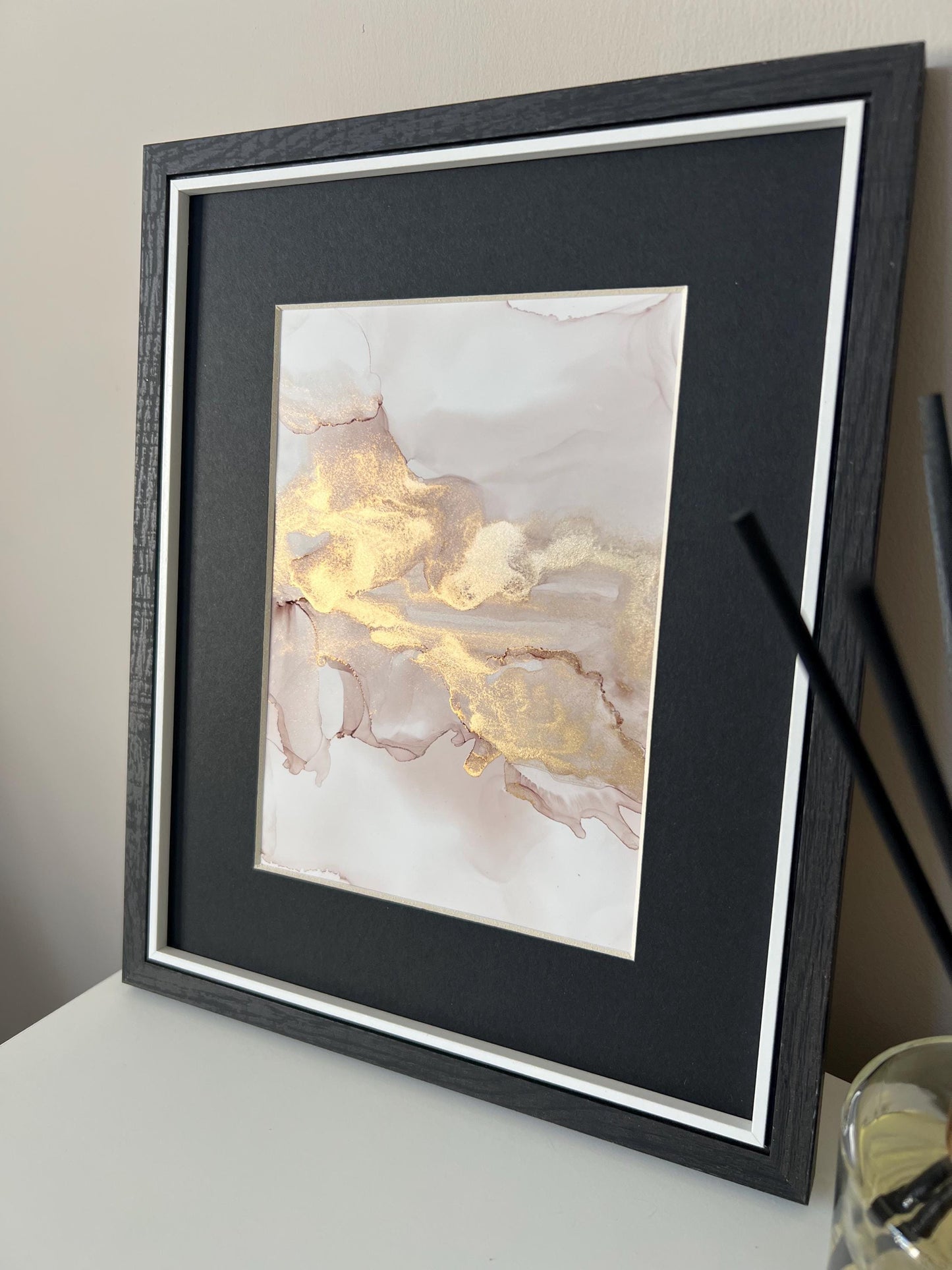 An A5 size contemporary ink painting with pink and gold metallic colors, presented in a black frame, placed on a white surface next to decorative items.