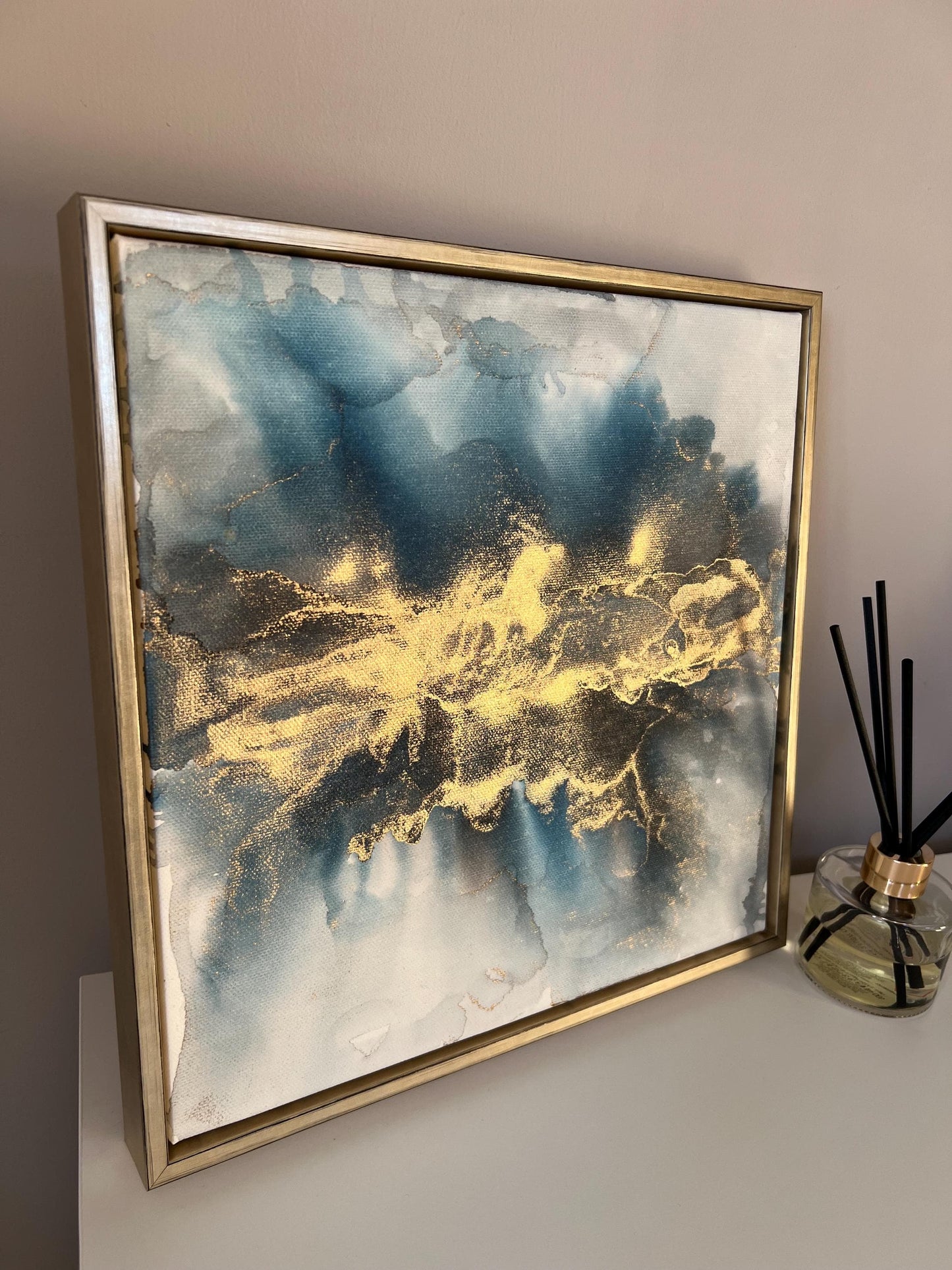 Side view of a one-off statement canvas painting featuring an abstract design in alcohol inks with gold and blue colours, with a metallic finish and in a gold finish box frame.