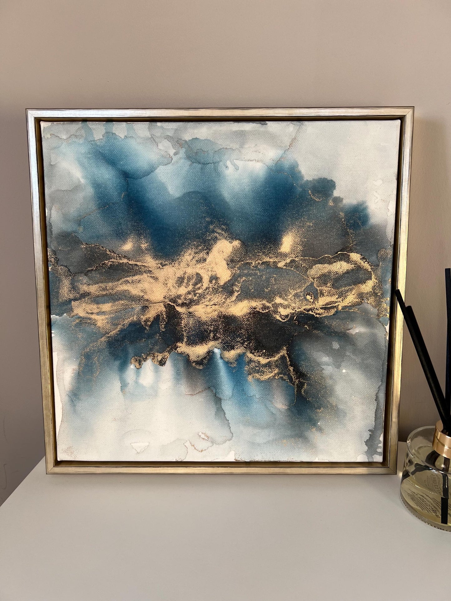 A one-off statement canvas painting featuring an abstract design in alcohol inks with gold and blue colours, with a metallic finish and in a gold finish box frame.