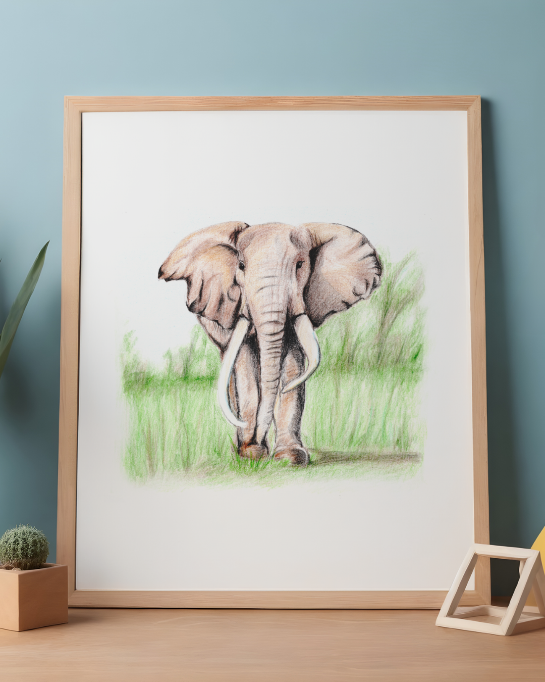 A framed art print featuring a colored pencil drawing of an elephant standing in green grass, displayed on a wooden table against a light blue wall with small decorative plants and a geometric stand.
