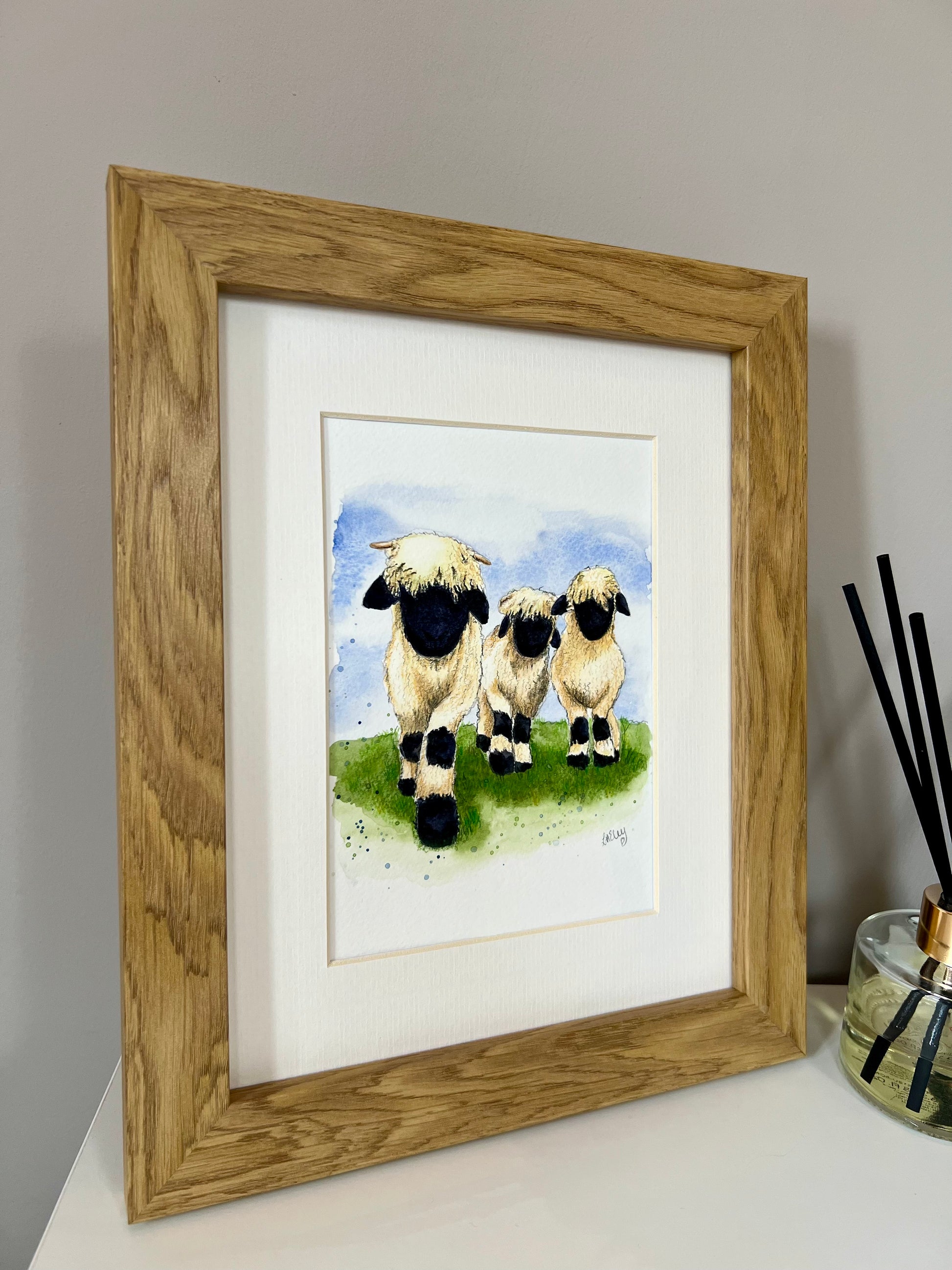 A framed watercolor painting of three Valais Blacknose sheep standing in a field, with a wooden oak finish frame and an ivory-toned mount.