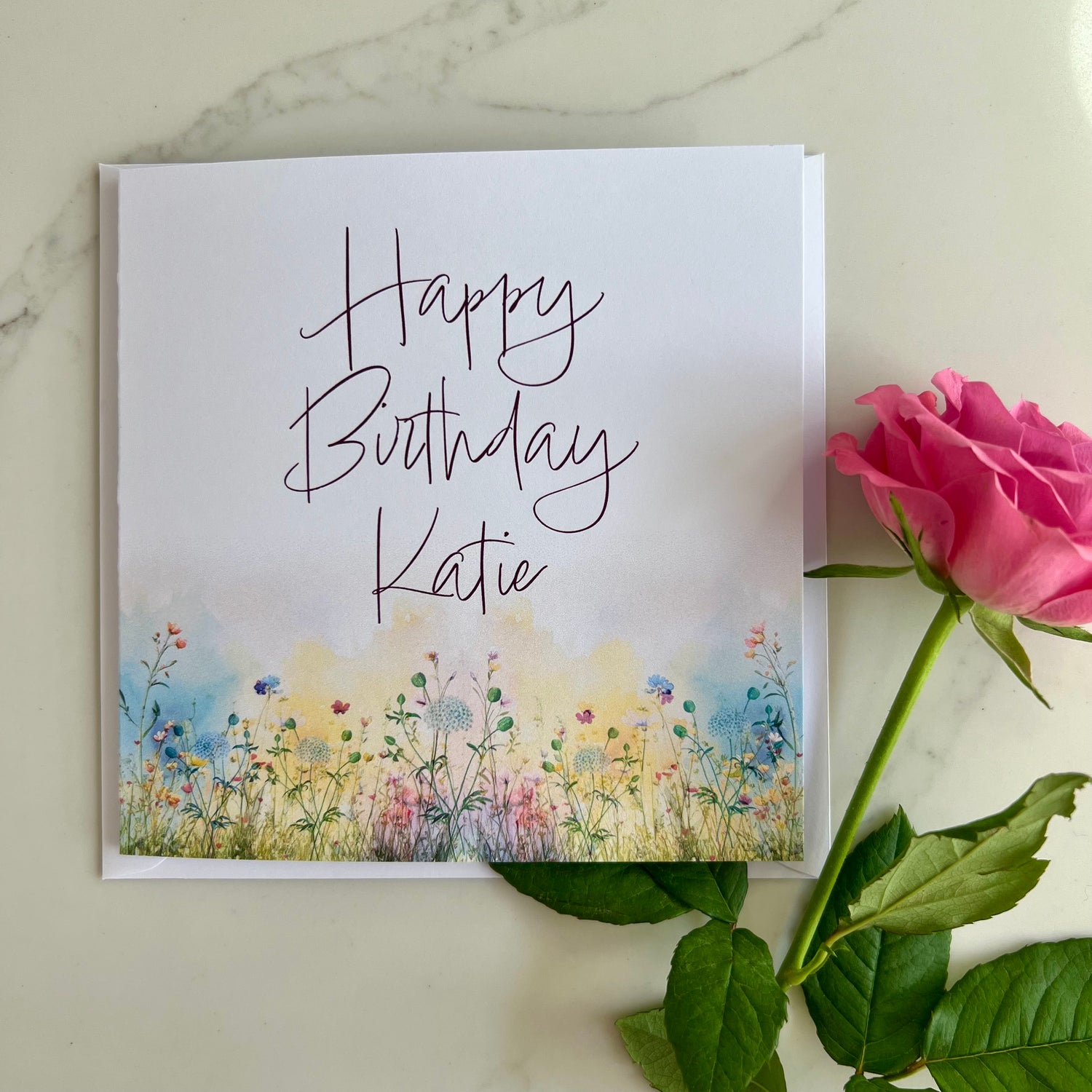 Personalised Greeting card with 'Happy Birthday Katie' text and floral design, accompanied by a pink rose on a marble surface.