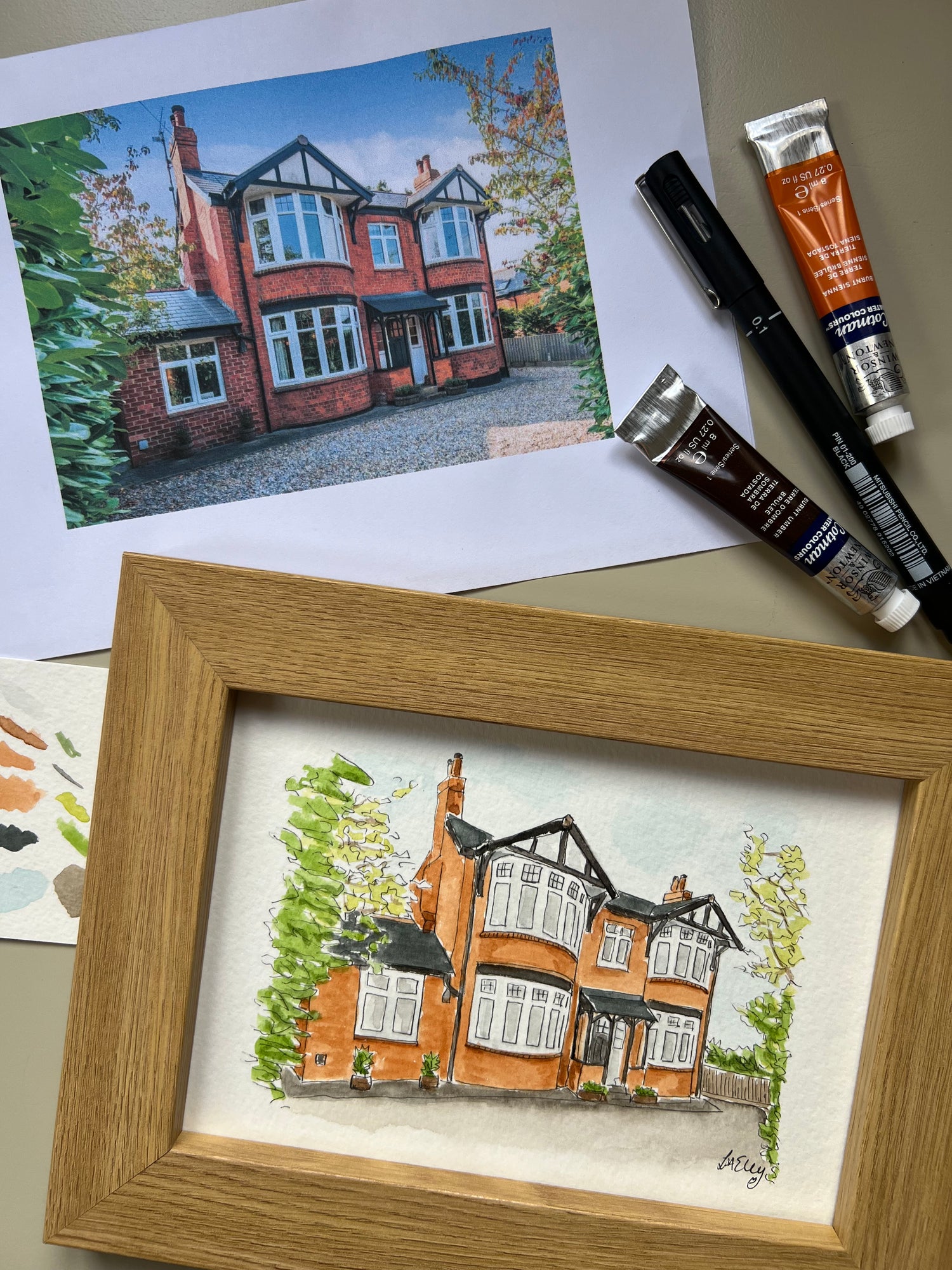 Framed artwork of a house portrait with paint tubes and original photo on paper in the background.