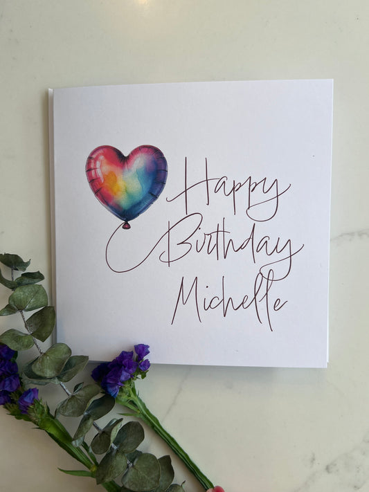 Birthday card with a heart-shaped balloon and personalized name on a marble surface with flowers.