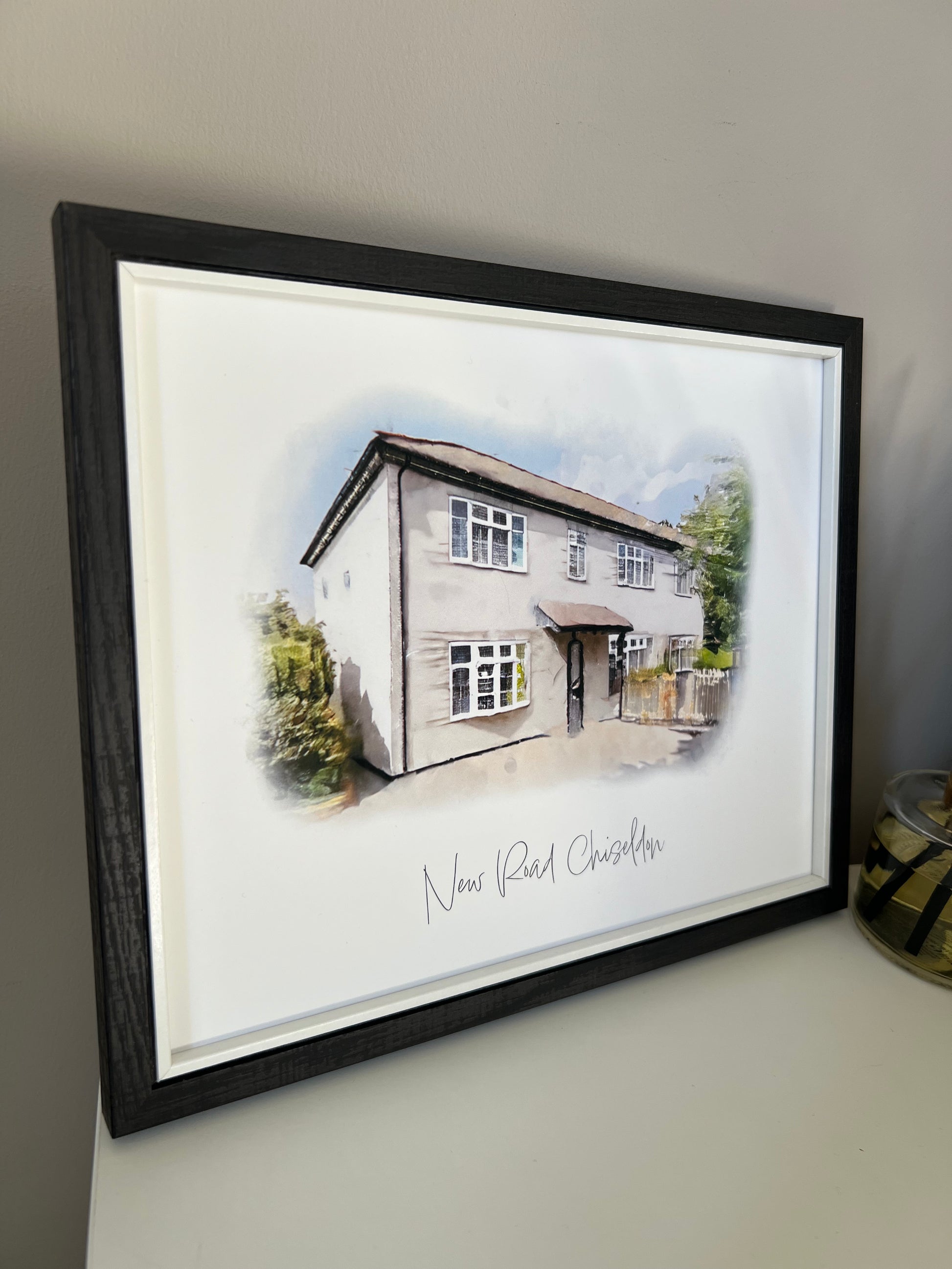 Side view of a framed artwork of a cottage house portrait in digital watercolour in a black frame.