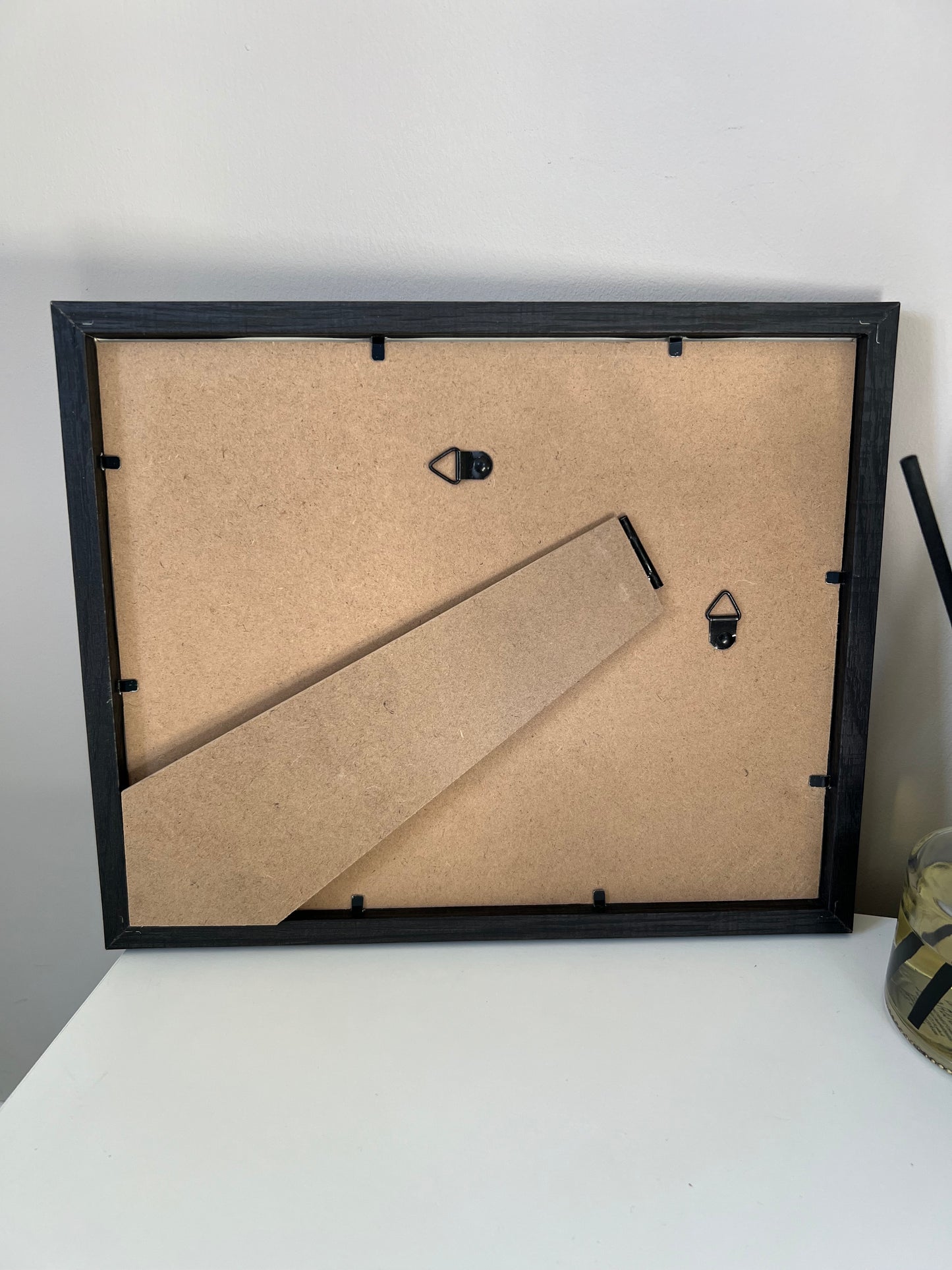 Black picture frame with mdf backing with back stand and hanging hooks on a white wall.