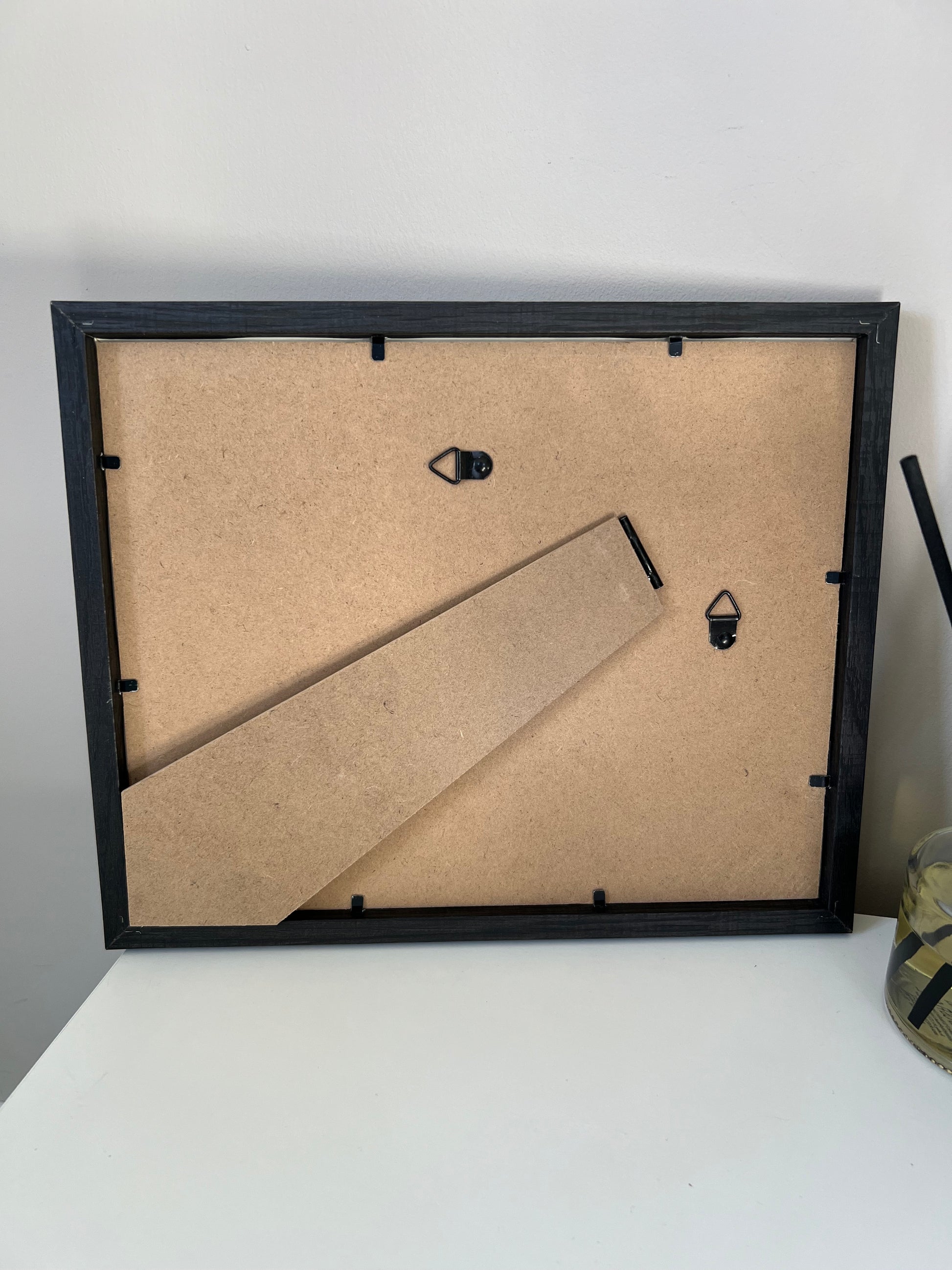 Black picture frame with mdf backing with back stand and hanging hooks on a white wall.