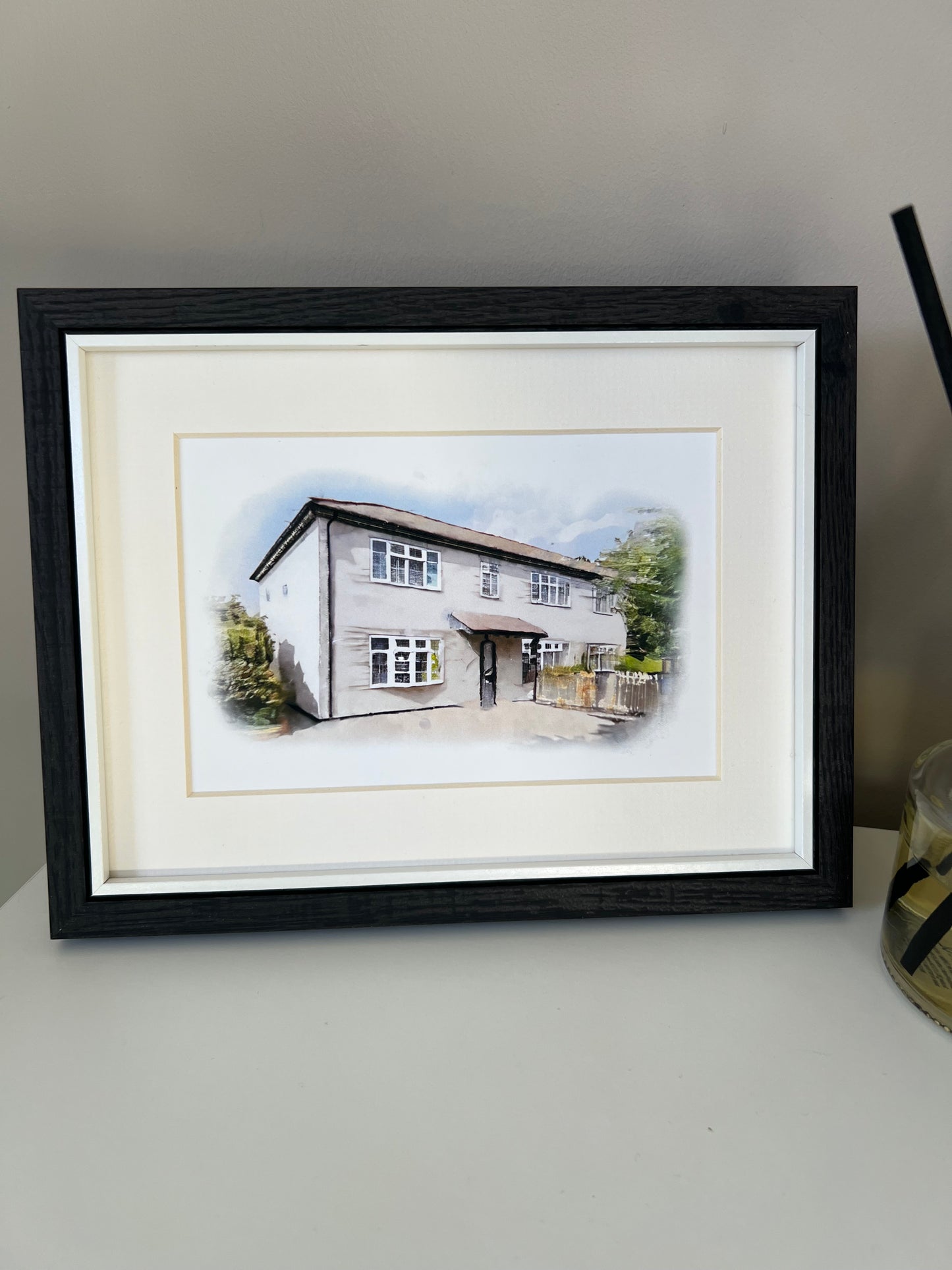 Framed artwork of a cottage house portrait in digital watercolour in a black frame and ivory mount. 