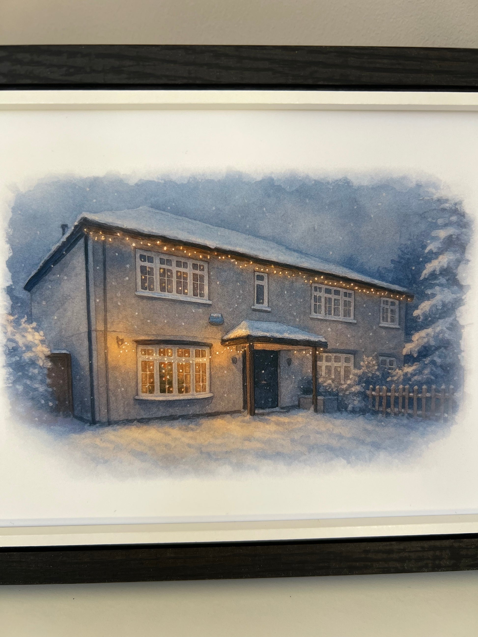 Close up of a framed artwork of a cottage style  house portrait in a winter snowy scene  in digital watercolour in a black finish frame.