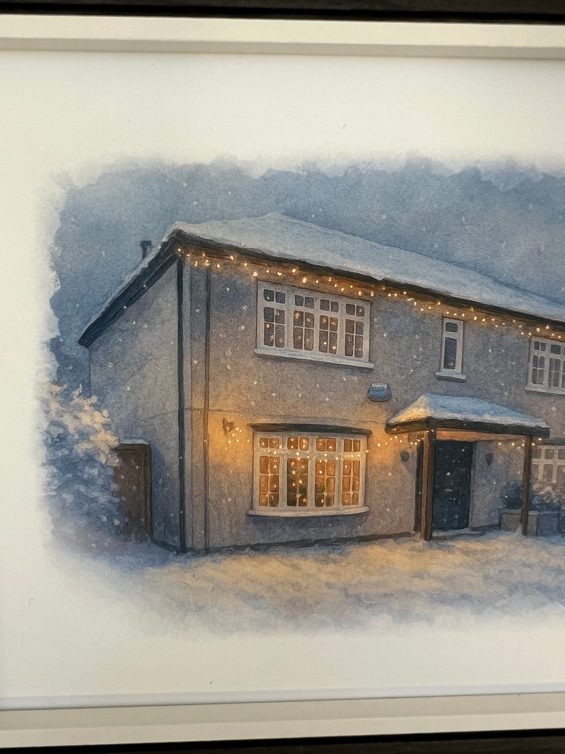 Close up of a framed artwork of a cottage style  house portrait in a winter snowy scene in digital watercolour in a black finish frame.