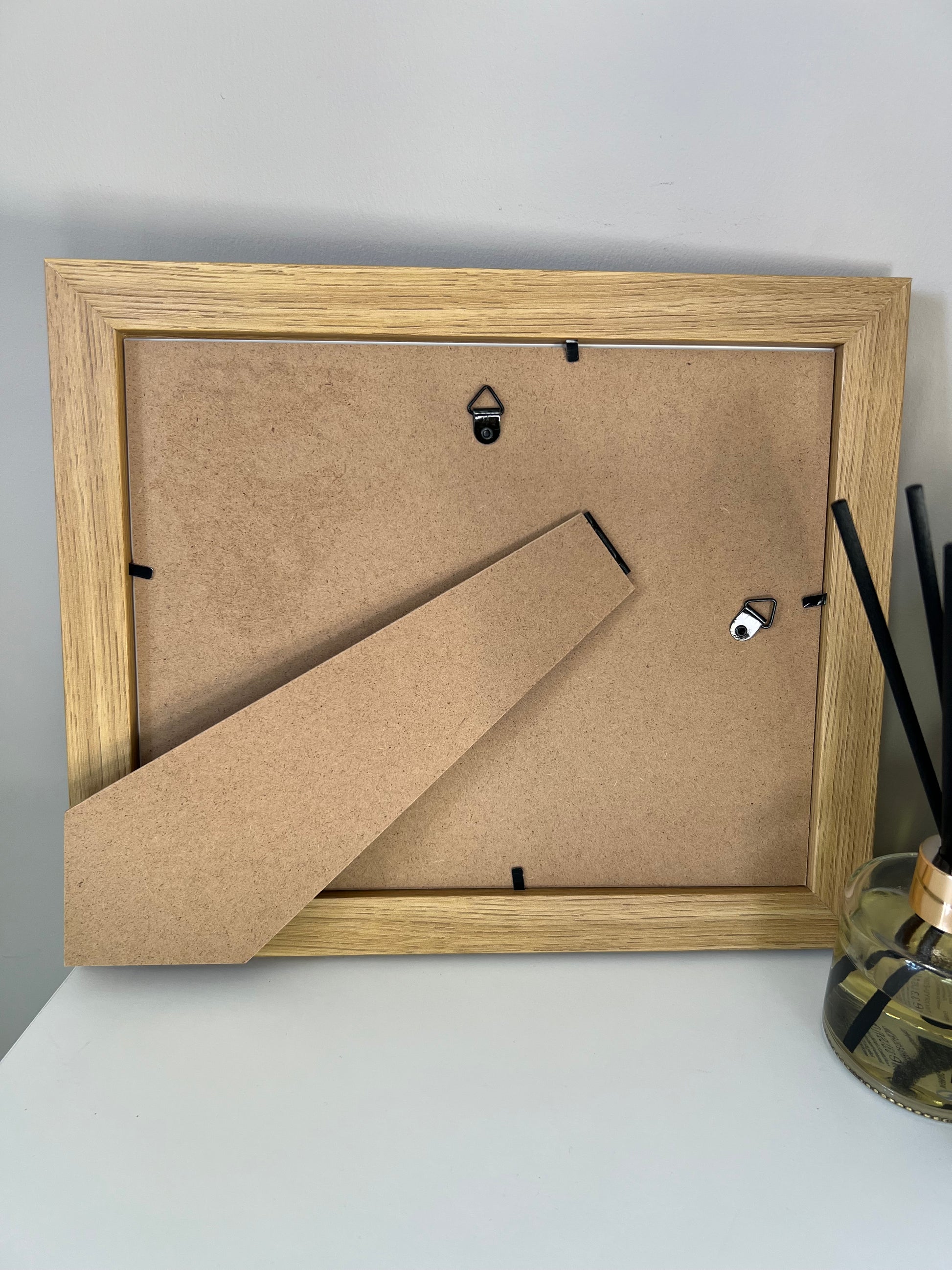 Oak finish picture frame with mdf backing with back stand and hanging hooks on a white wall.