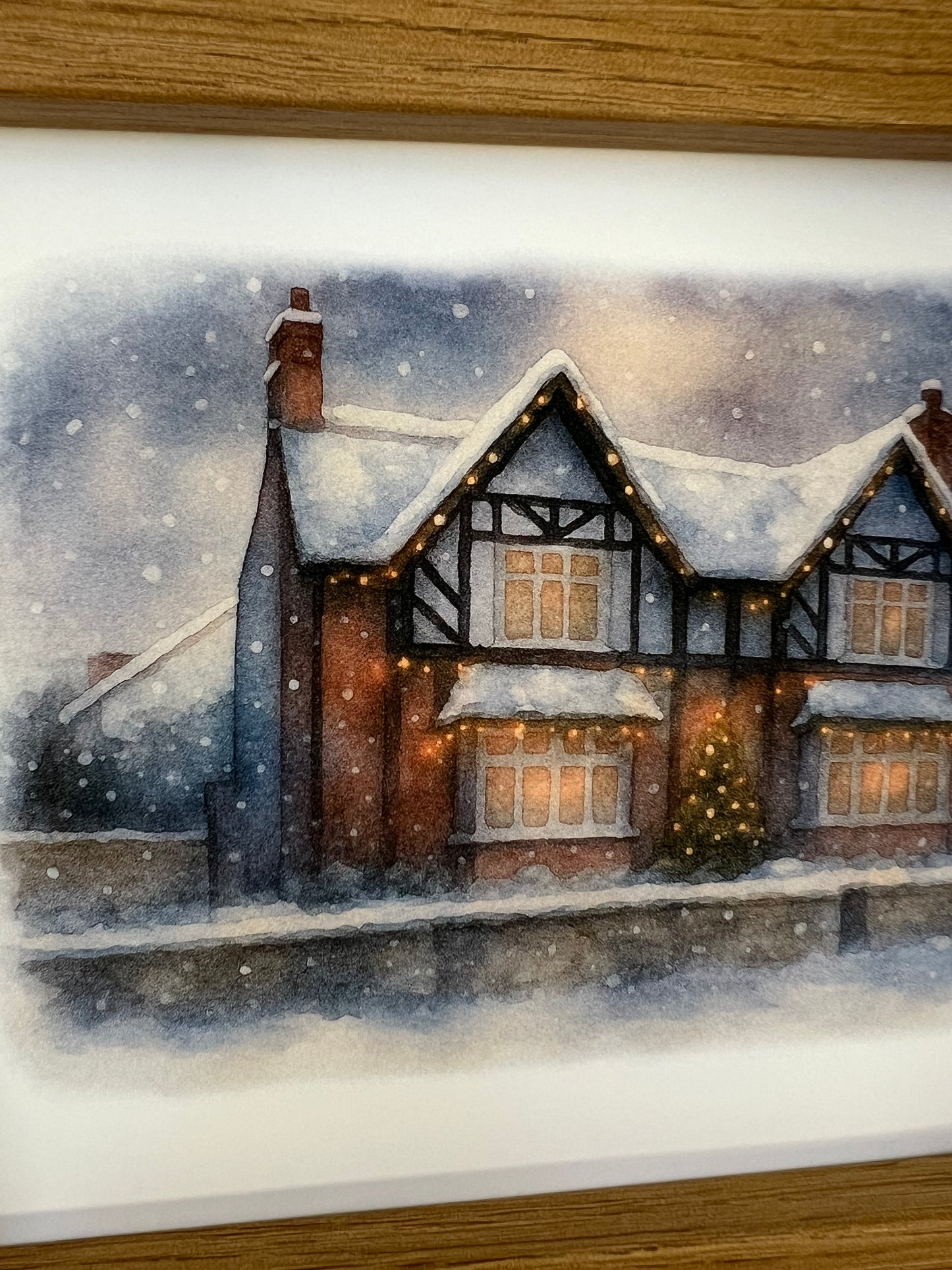 Close up of a framed artwork of a Tudor style period house portrait in a winter snowy scene  in digital watercolour in an oak finish frame.