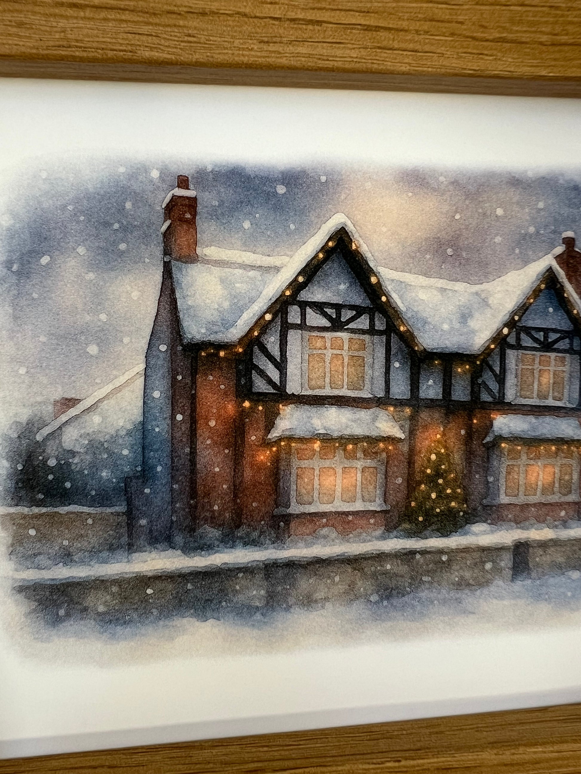 Close up of a framed artwork of a Tudor style period house portrait in a winter snowy scene  in digital watercolour in an oak finish frame.