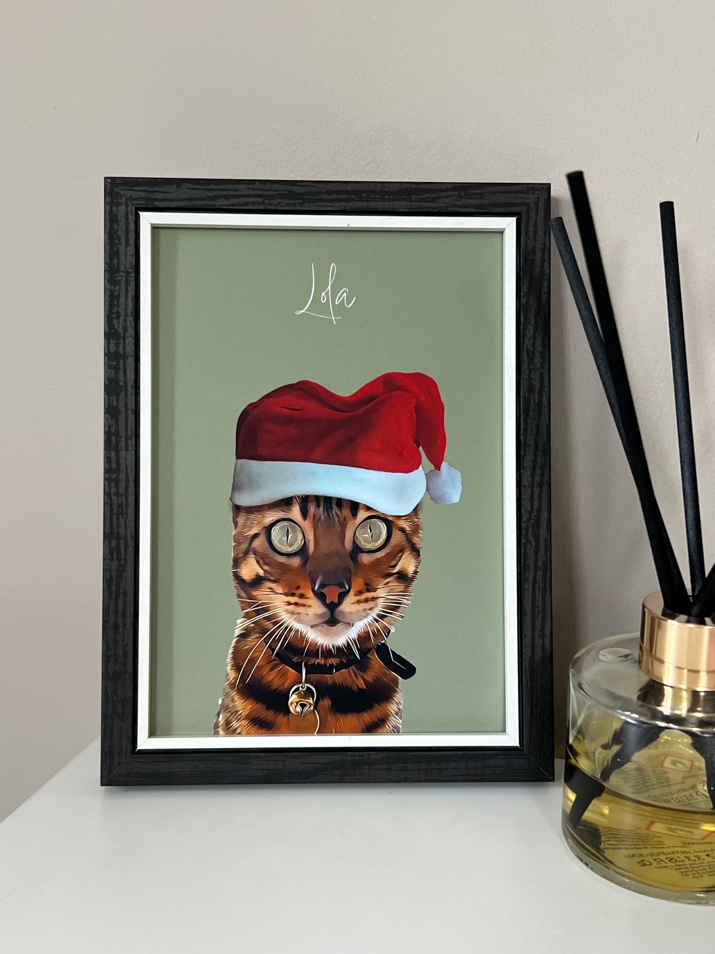 Framed artwork of a Bengal Cat wearing a Santa hat in a black frame on a beige wall.